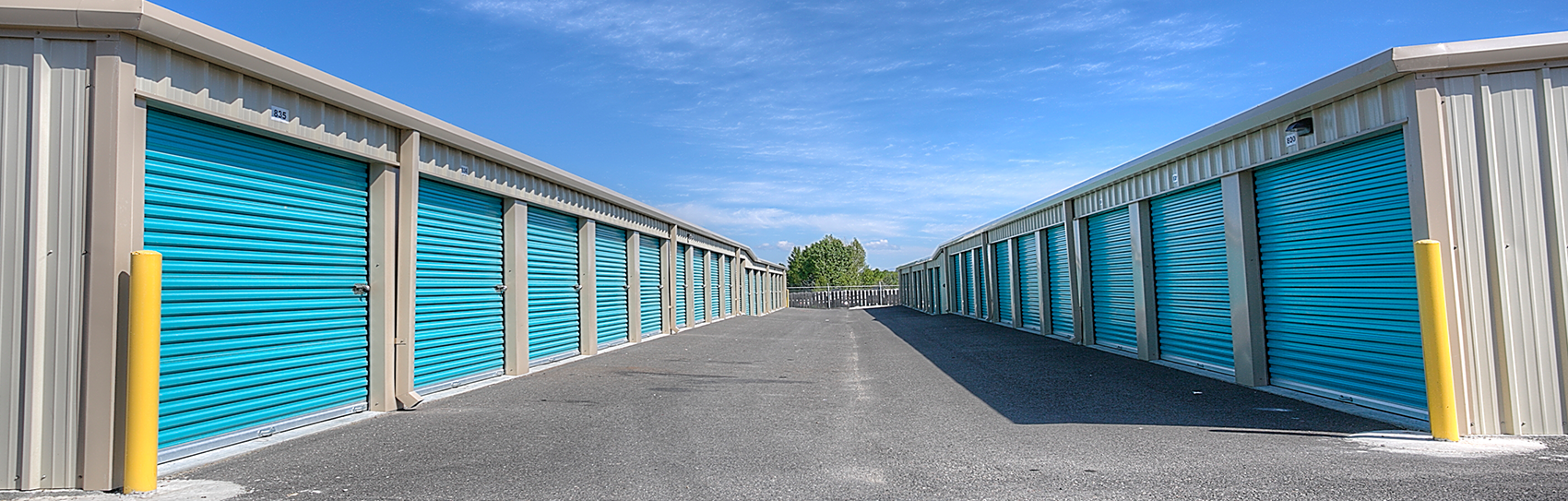 Rows of blue storage unit doors lining both sides of a gravel driveway under a clear sky