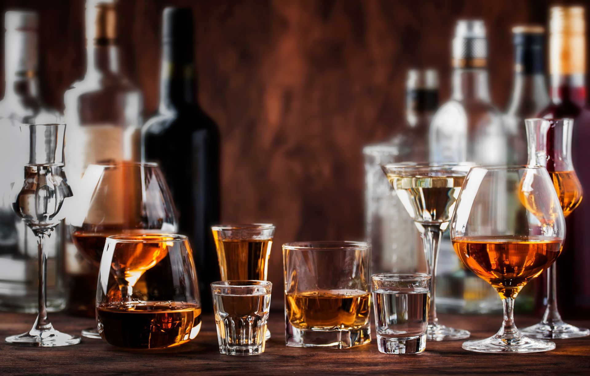 Verschiedene Whiskyflaschen und -gläser auf einer Holzbar, mit bernsteinfarbenem Whisky und warmen Spiegelungen.