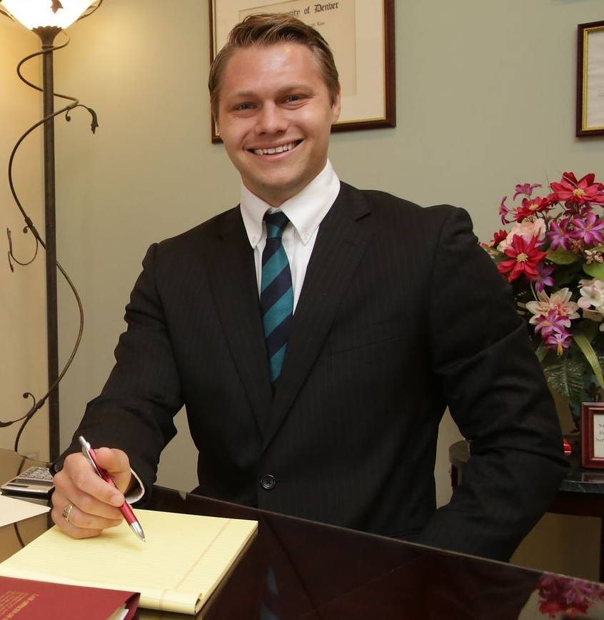 Smiling professional in a suit at a desk, holding a pen over a notebook in an office.