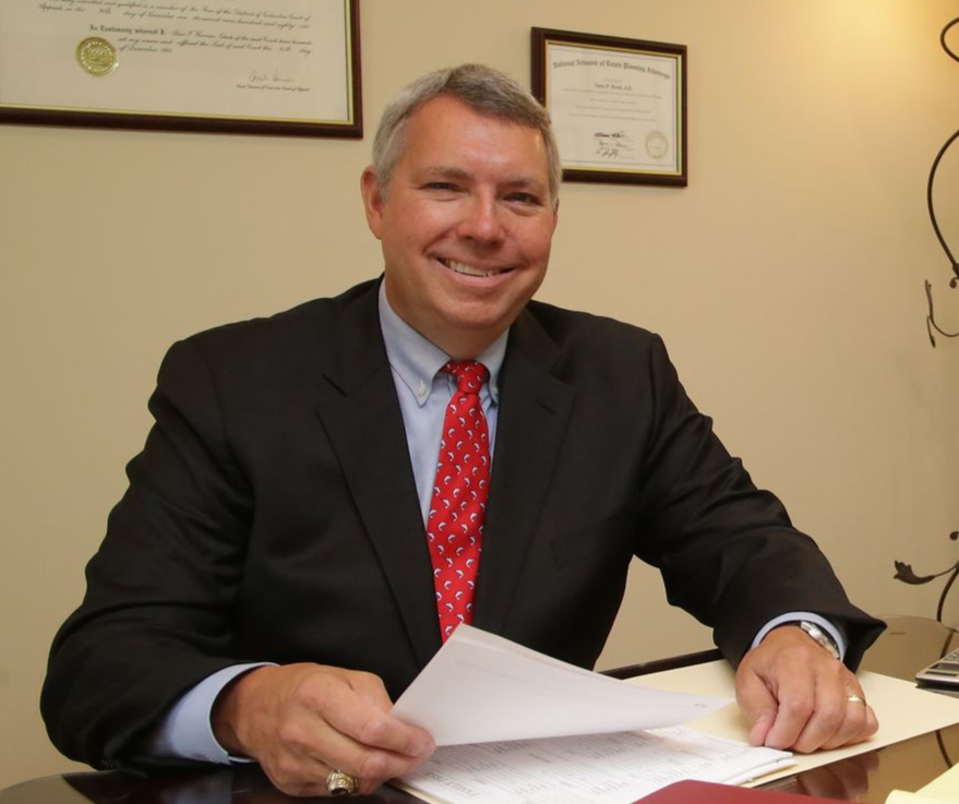 Smiling man in a suit and red tie seated at a desk with papers in an office