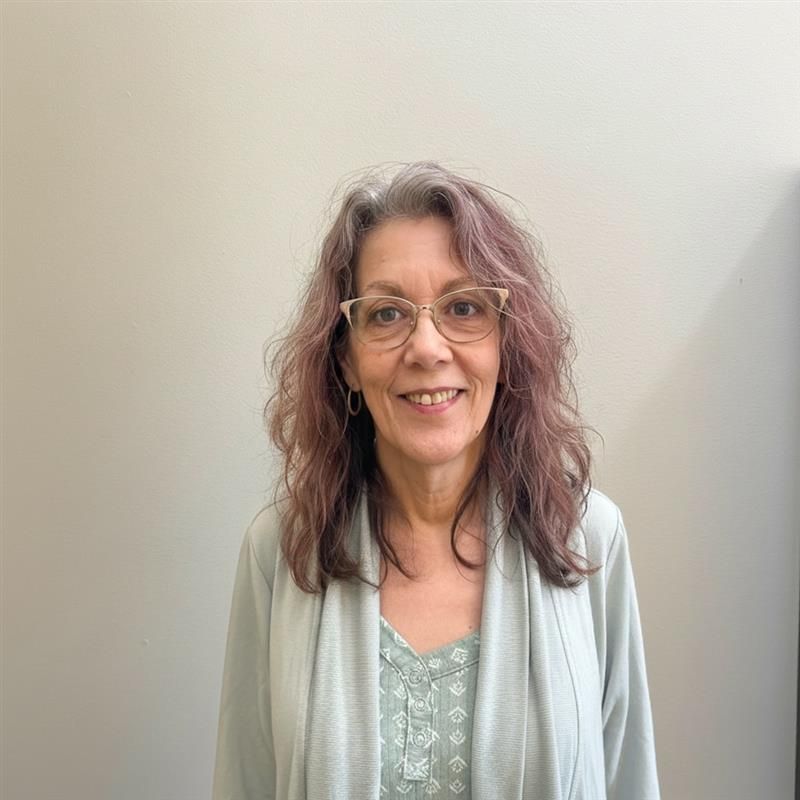 Smiling person with wavy hair and glasses, wearing a light cardigan against a plain beige wall.
