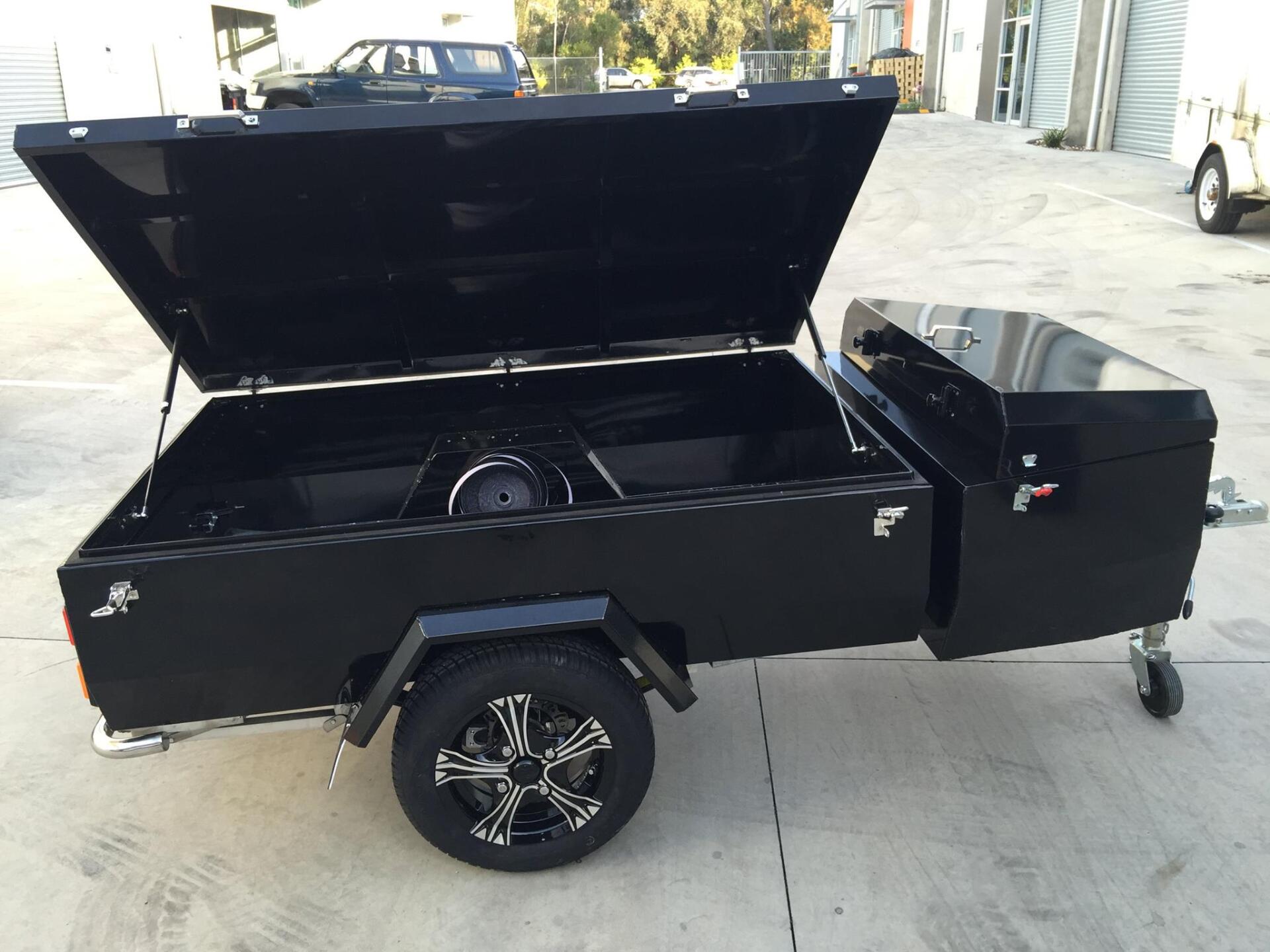 Black utility trailer with an open lid, spare tire, and wheels. The trailer is parked outside.