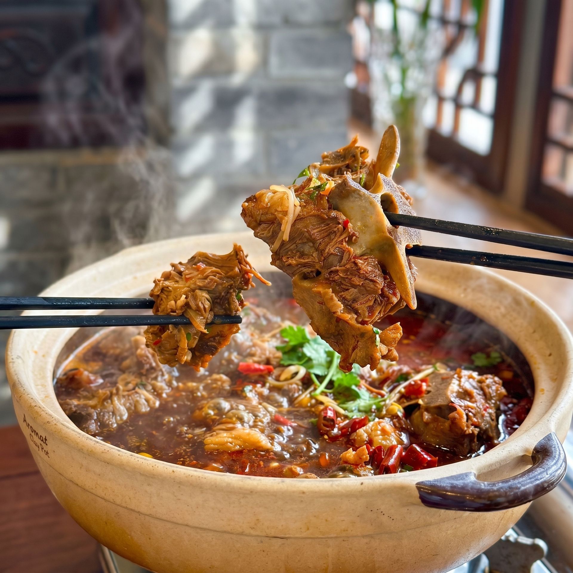 Chopsticks lifting meat from a spicy stew in a beige clay pot on a table by a window