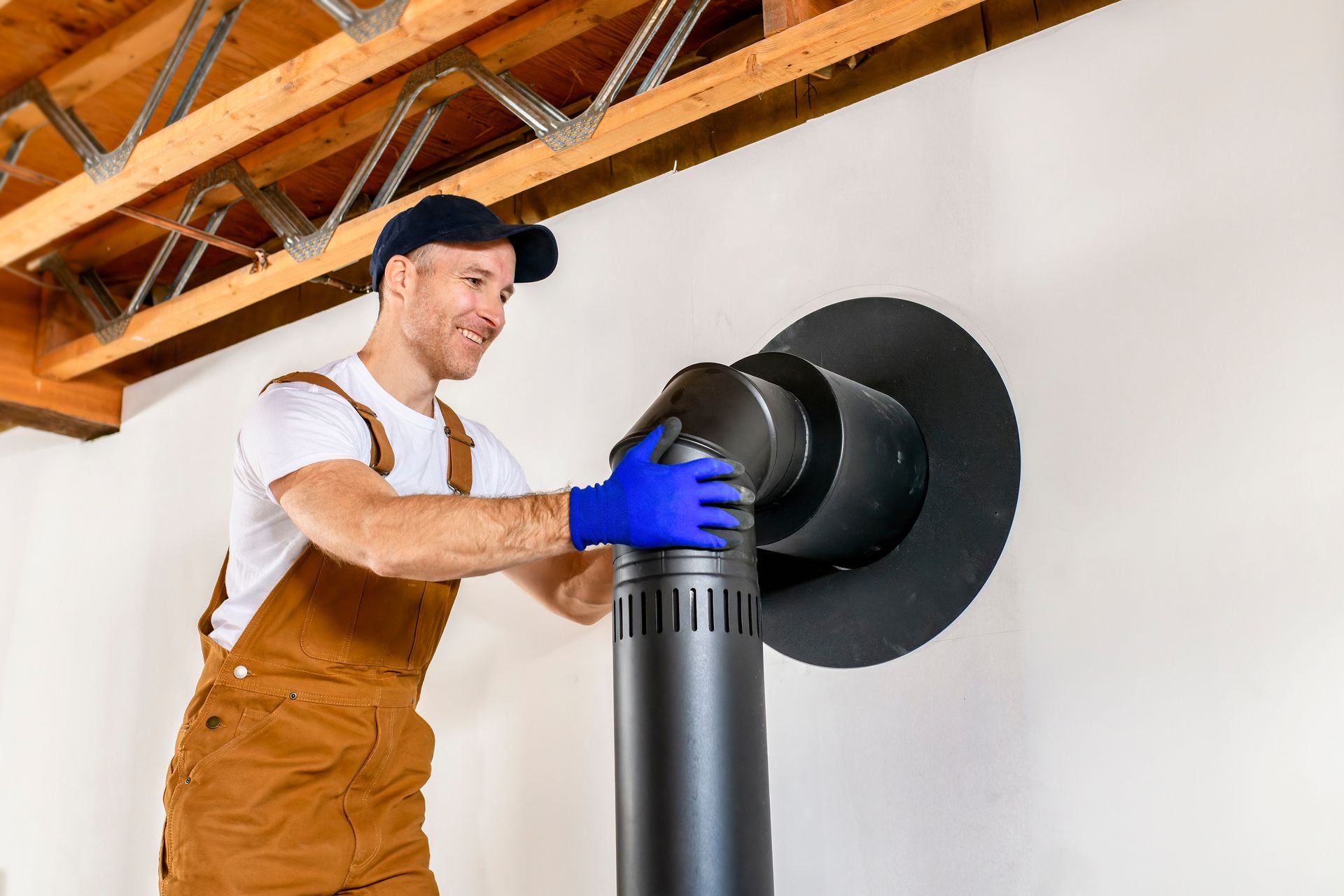 Man in overalls, blue gloves, installing black stove pipe into a wall vent.