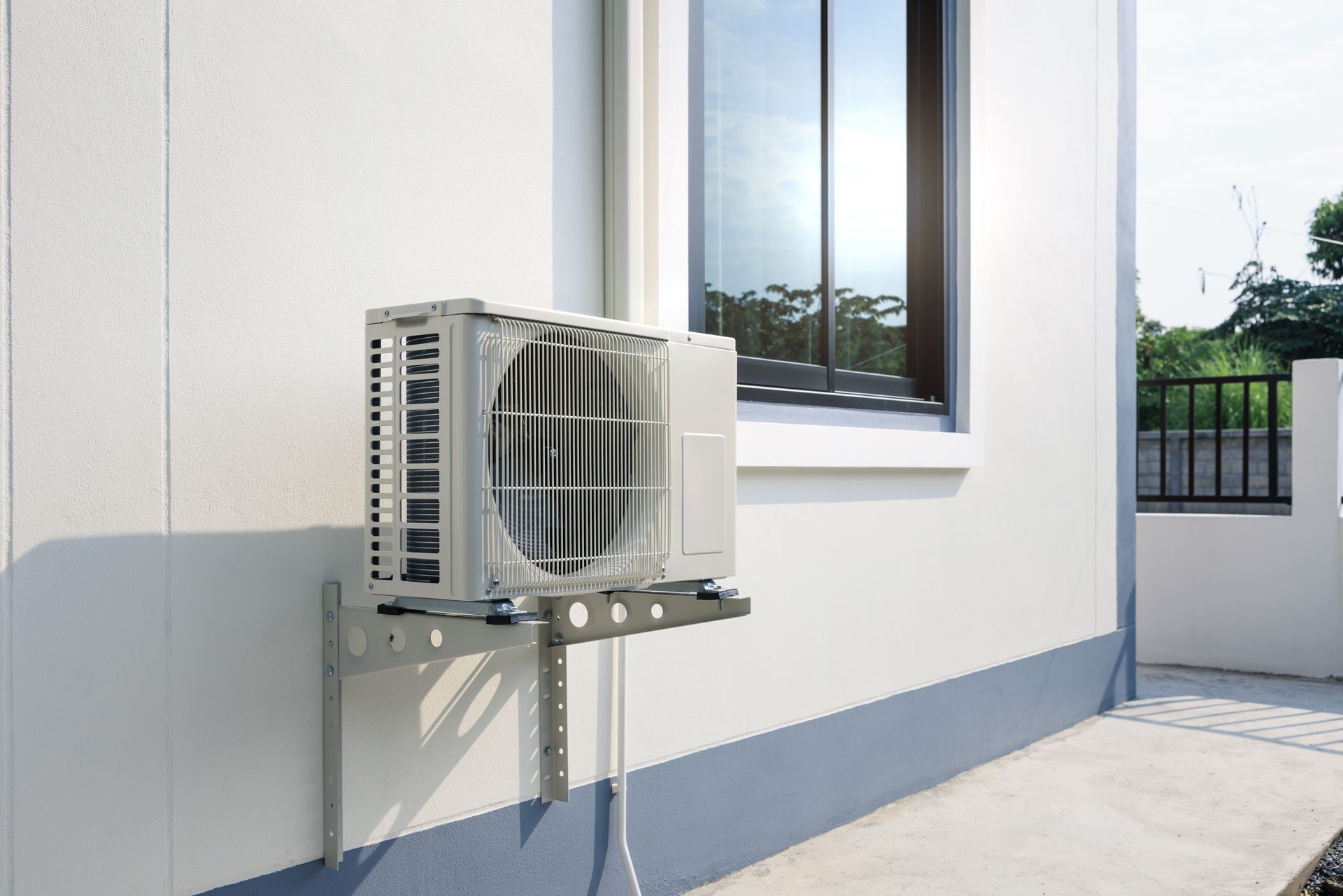 Exterior AC unit mounted on a white house wall near a window and a walkway.