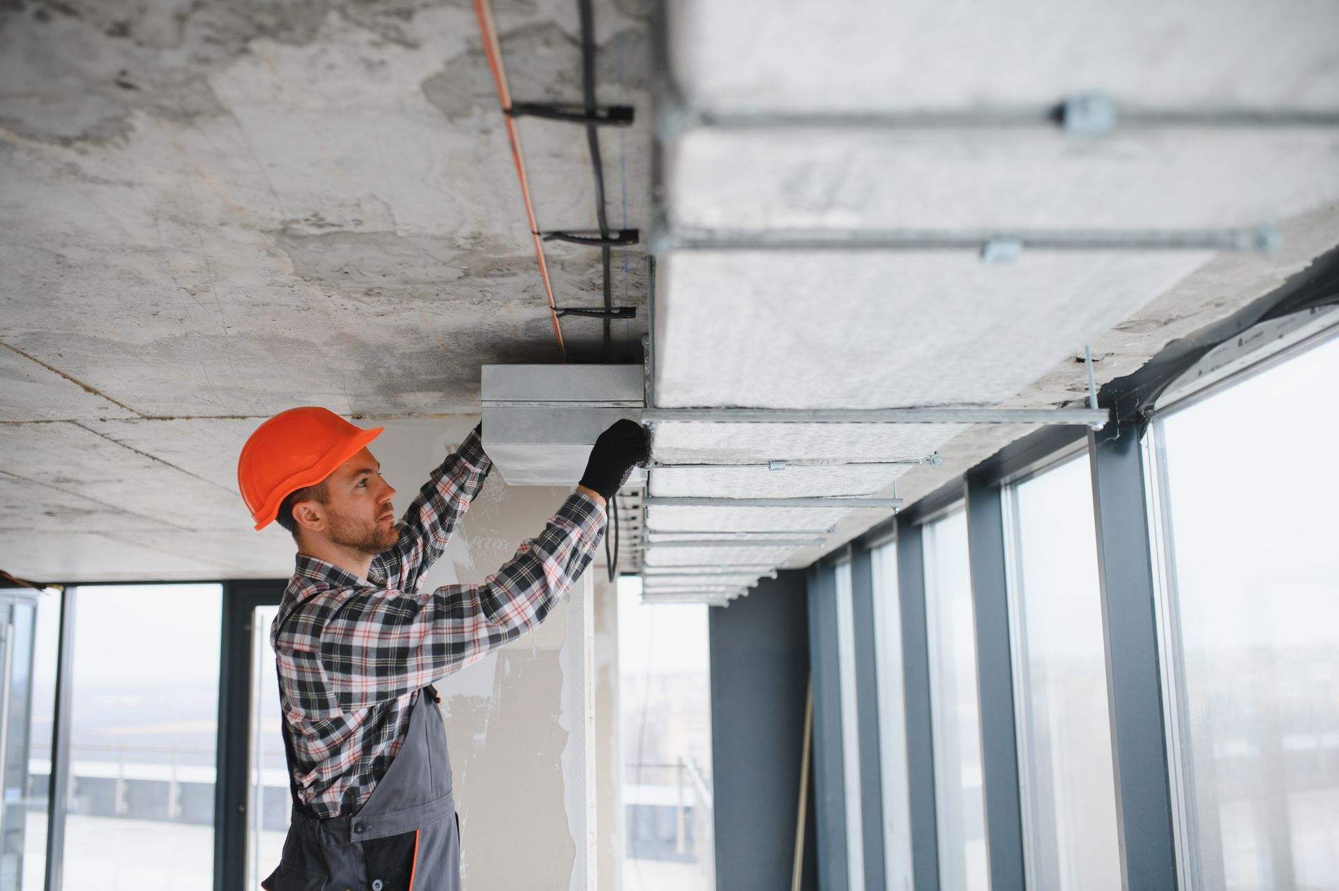 Construction worker in hard hat installs ceiling electrical components near large windows.
