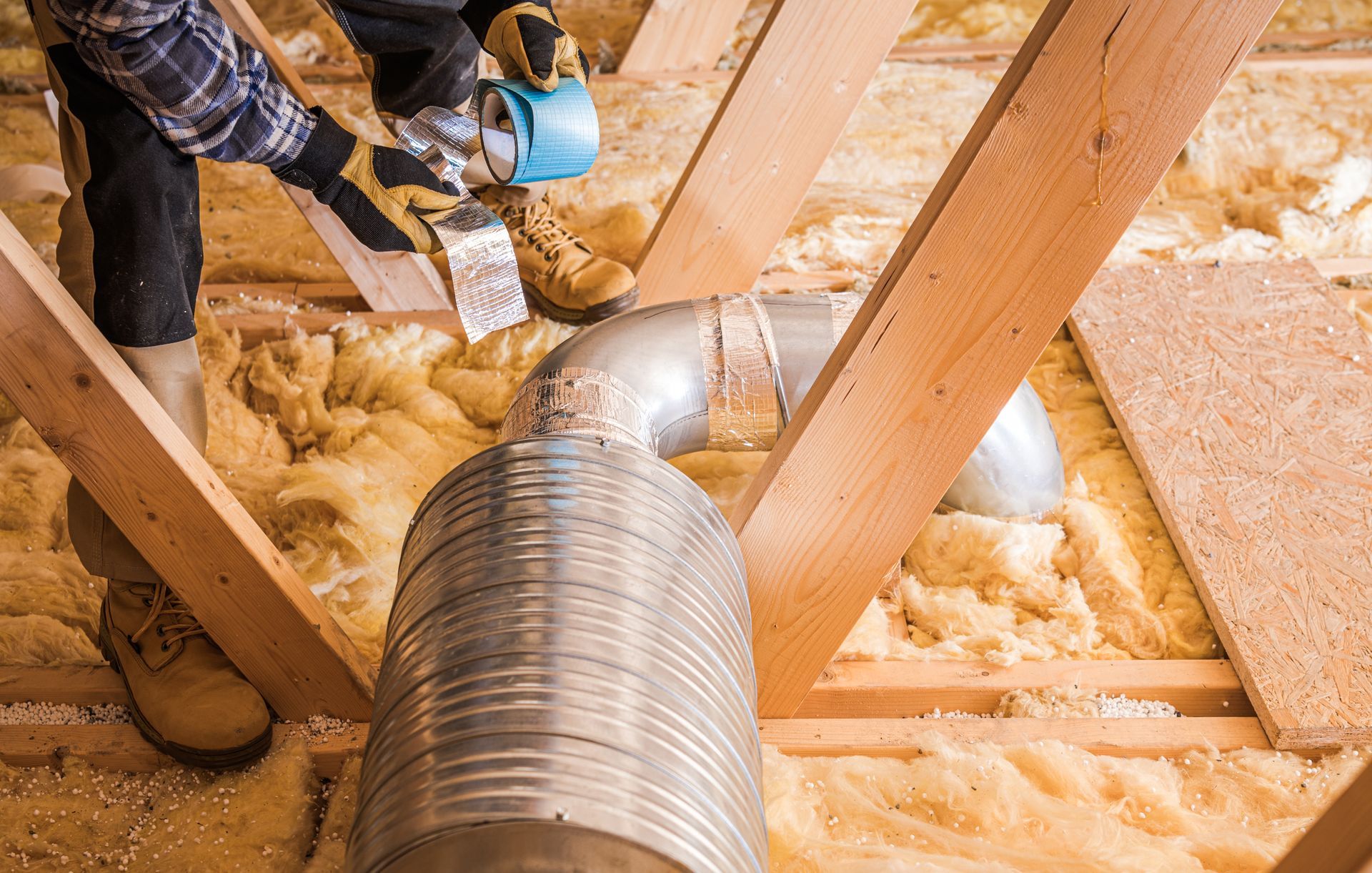 Person installing HVAC ductwork in an attic, surrounded by insulation and wooden beams.