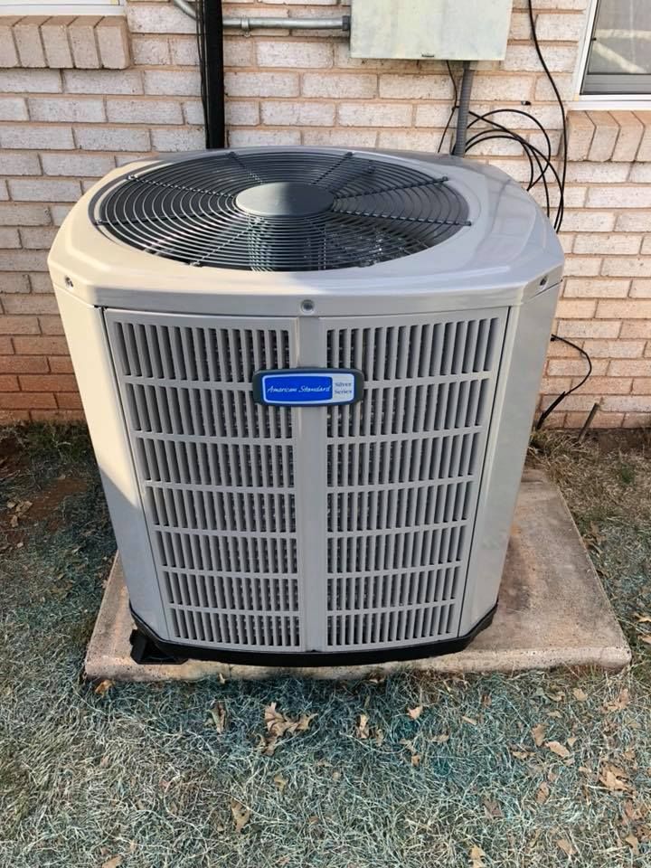 Gray air conditioning unit outside, sitting on a concrete pad. Brick wall background.