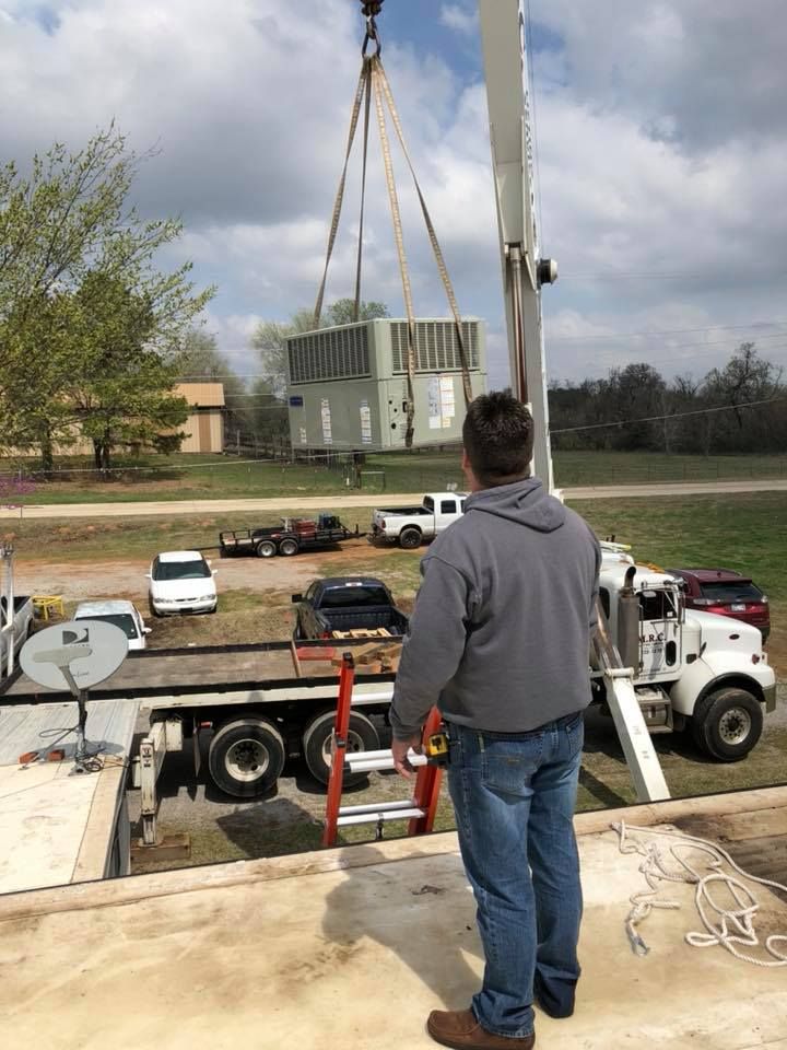 Person watches as a crane lifts an HVAC unit. Truck, vehicles, and trees are in the background.