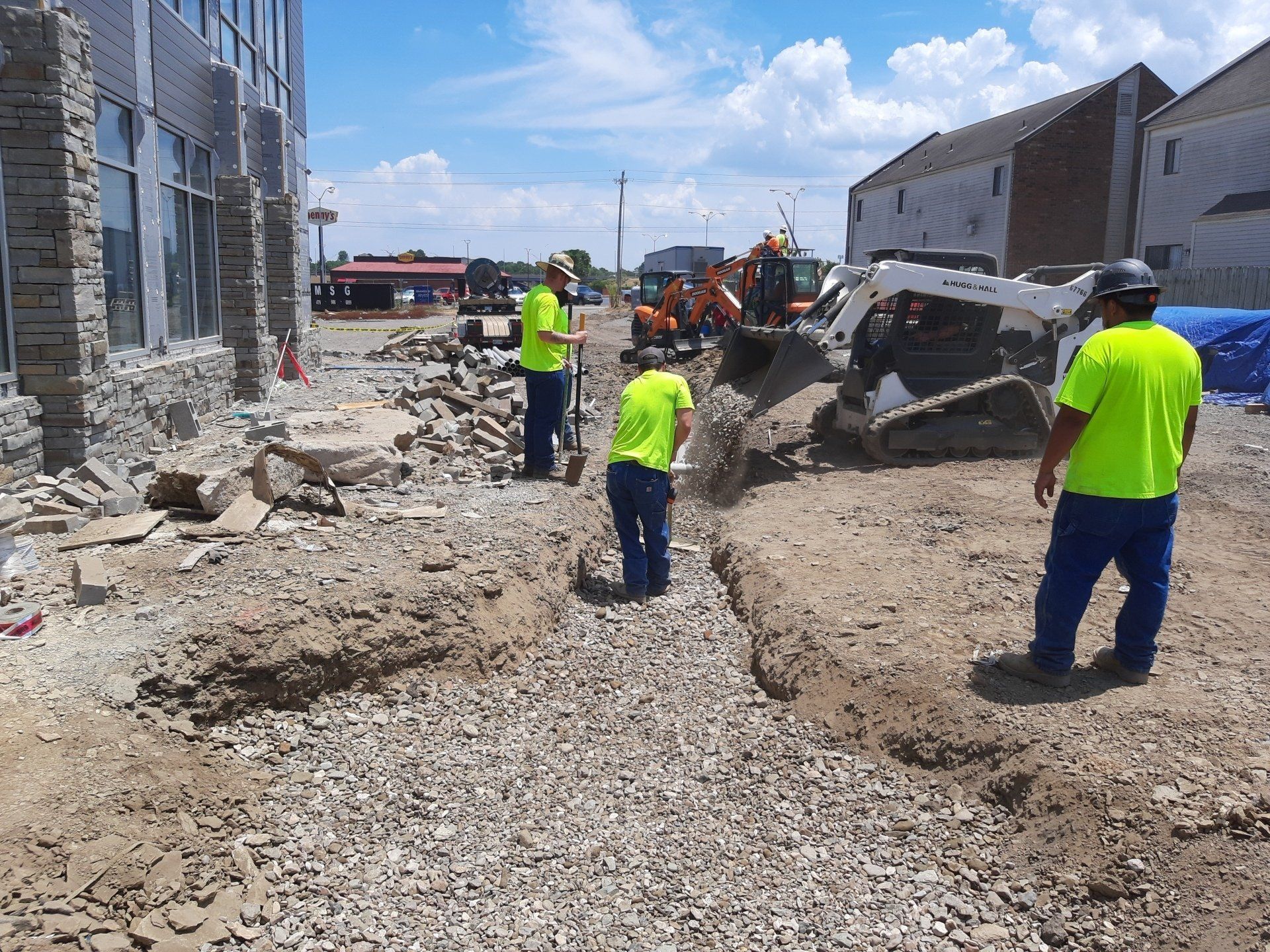 Construction workers in neon shirts digging near a building with heavy machinery on a sunny day.