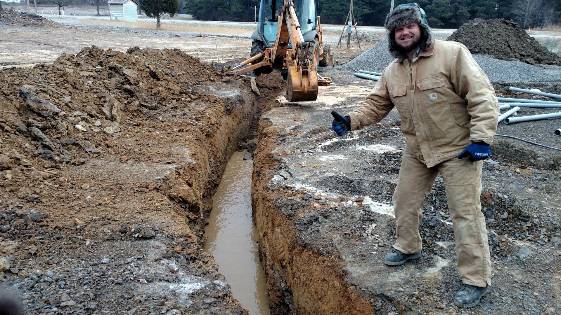 Man in work clothes points at trench dug by a backhoe on a construction site.