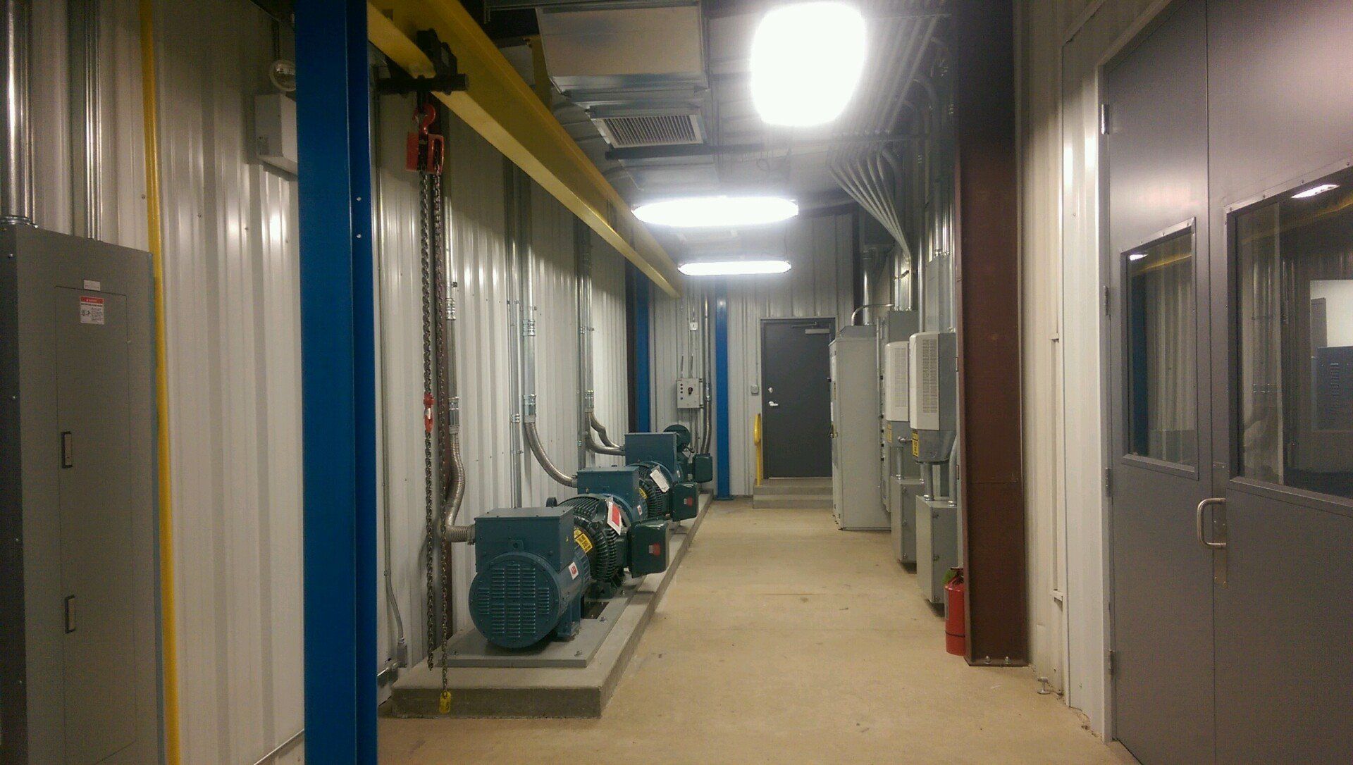 A narrow room with industrial equipment, including generators, lit by overhead lights.