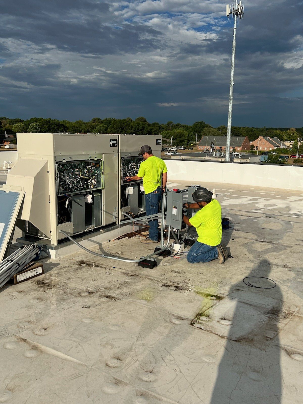 Two workers in safety vests on a rooftop, repairing HVAC units. Overcast sky.
