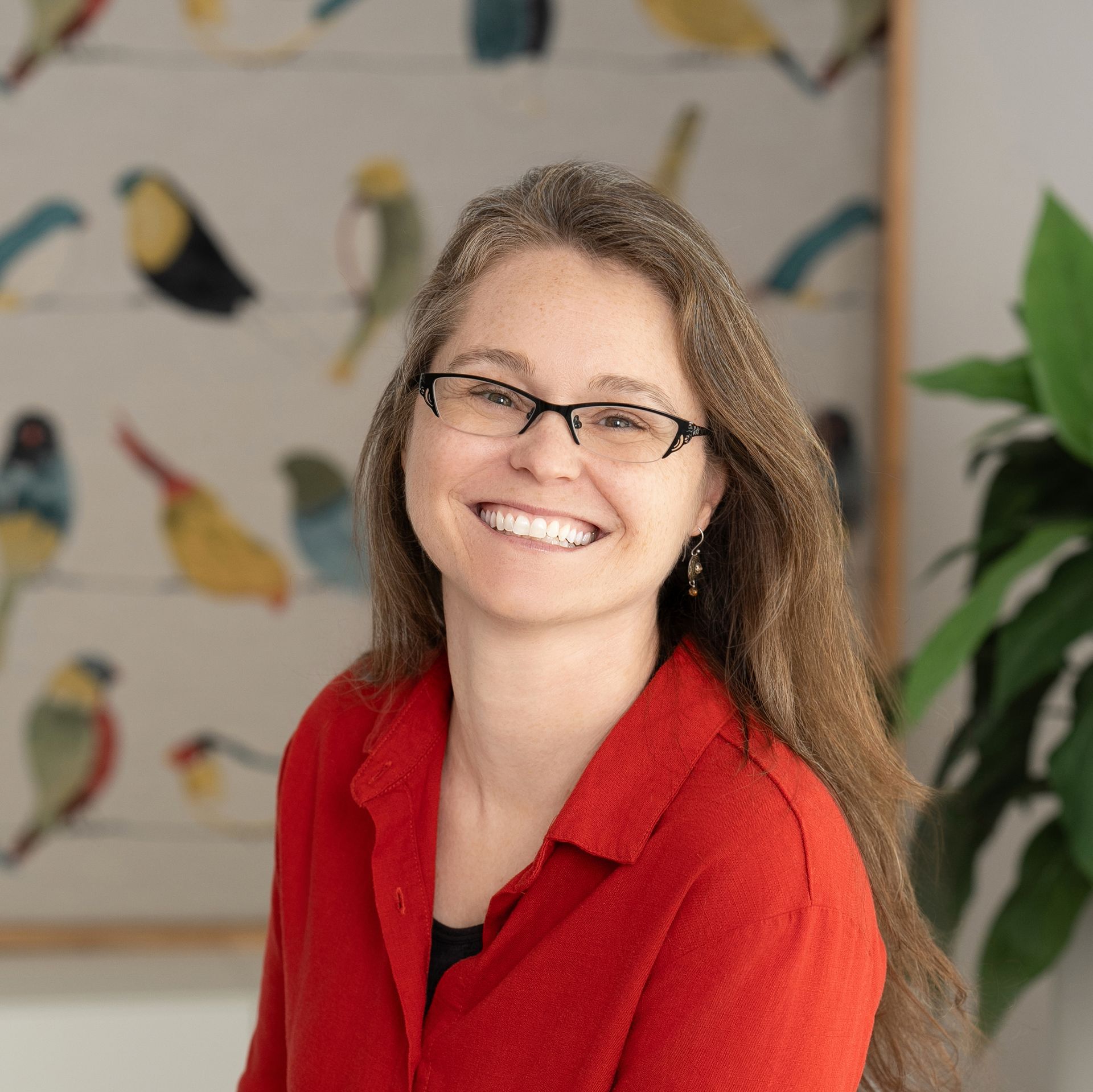 A woman wearing glasses and a red shirt is smiling in front of a painting of birds.