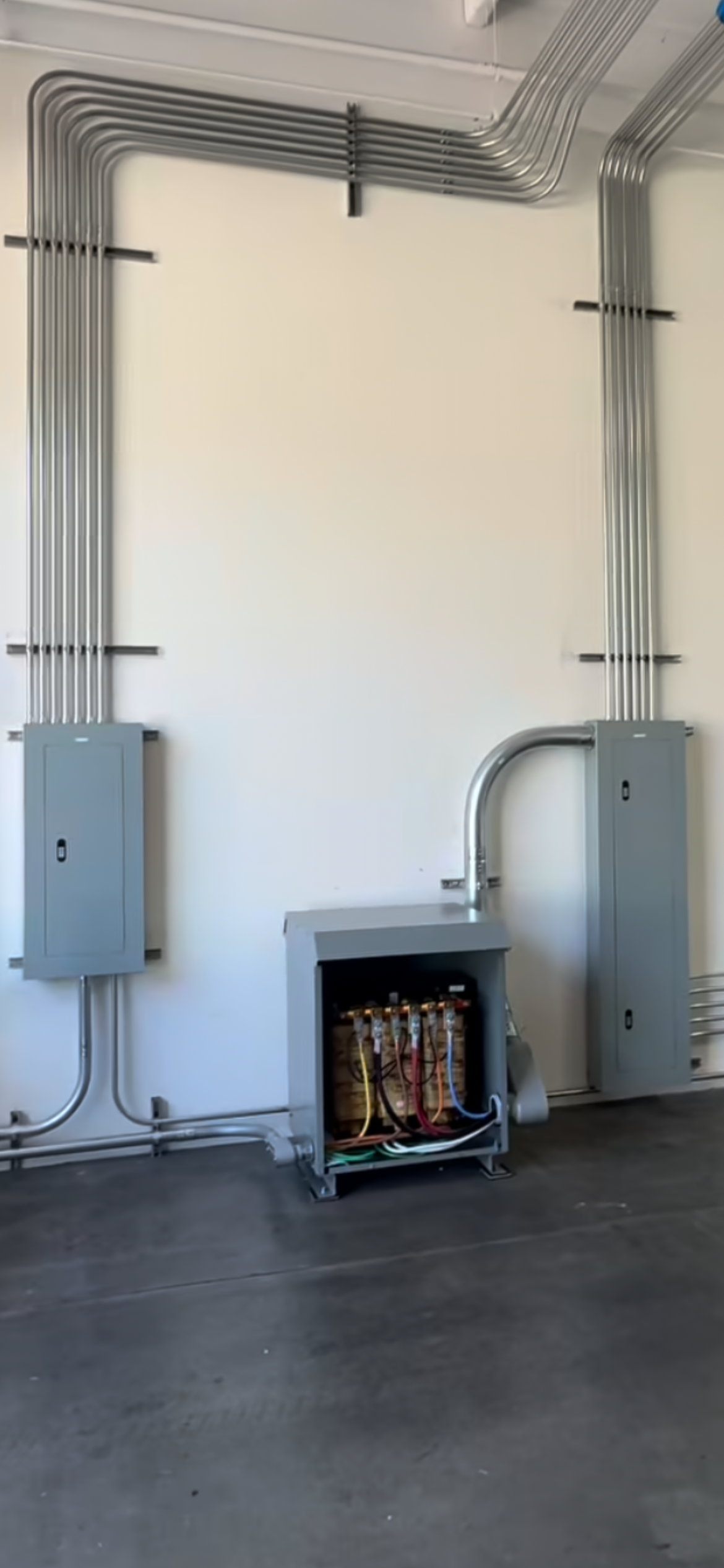 Electrical conduit and panels mounted on a white wall in a room with a concrete floor.
