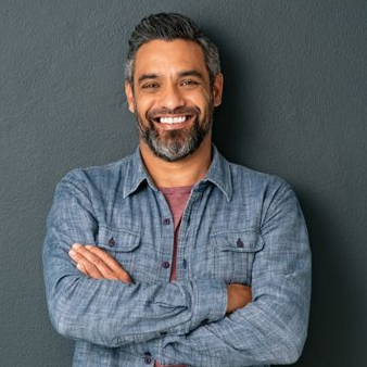Man with salt-and-pepper beard smiles, arms crossed, wearing blue button-up shirt over red tee, against gray wall.