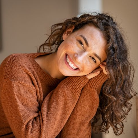 Woman with curly hair in a brown sweater smiles, leaning on her arms.