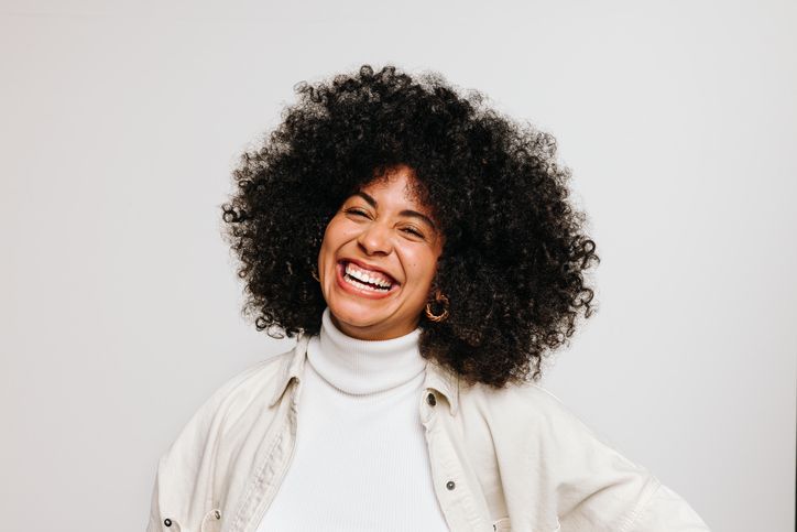 Woman with large afro smiling widely, wearing a white turtleneck and button-down shirt.