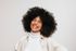 Woman with voluminous curly hair laughs, wearing a white turtleneck and light jacket against a white background.