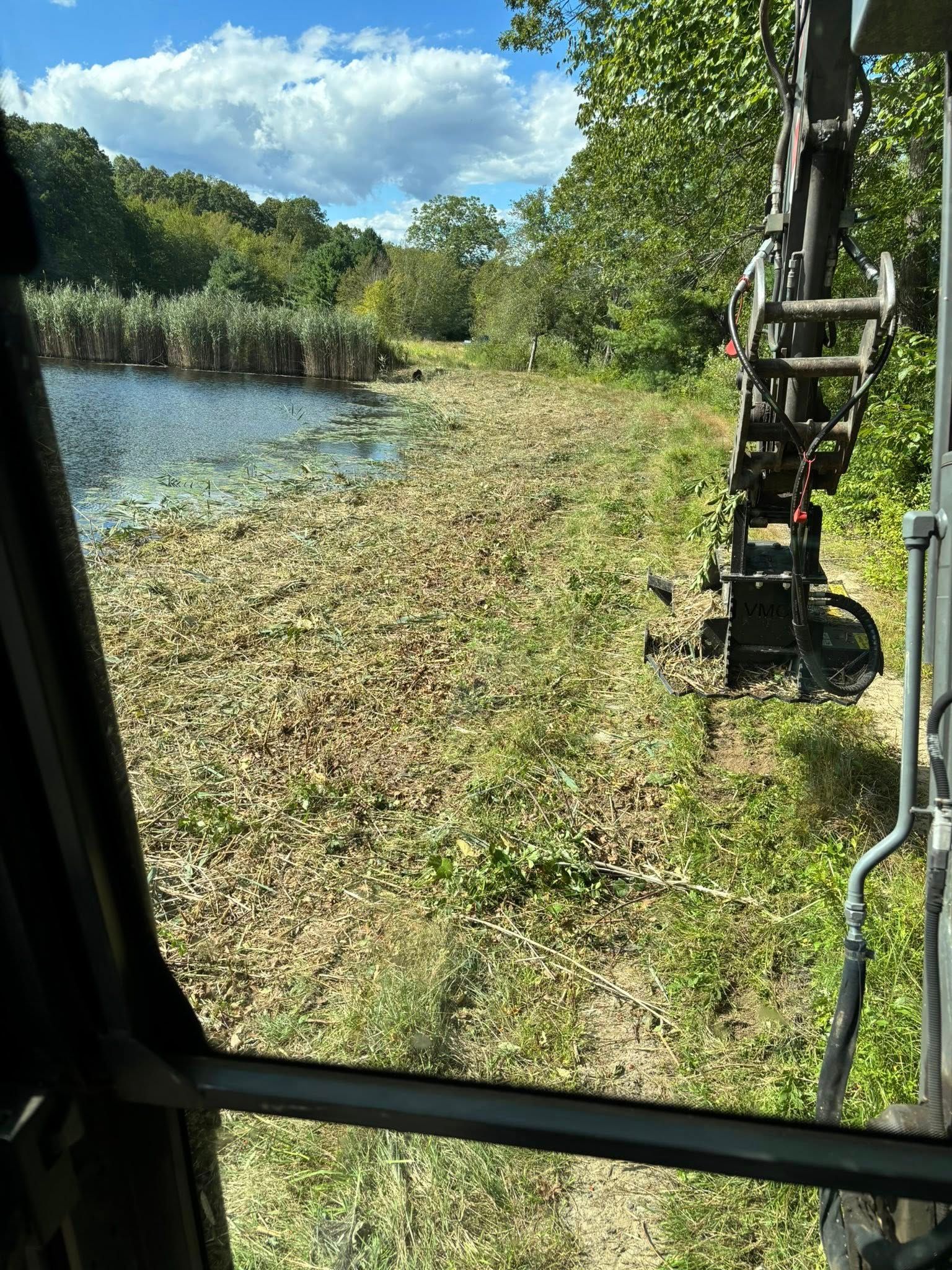 A machine clearing vegetation from the edge of a pond under a cloudy sky.