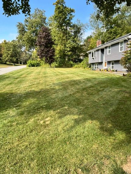 Green lawn in front of a gray house, surrounded by trees under a blue sky.
