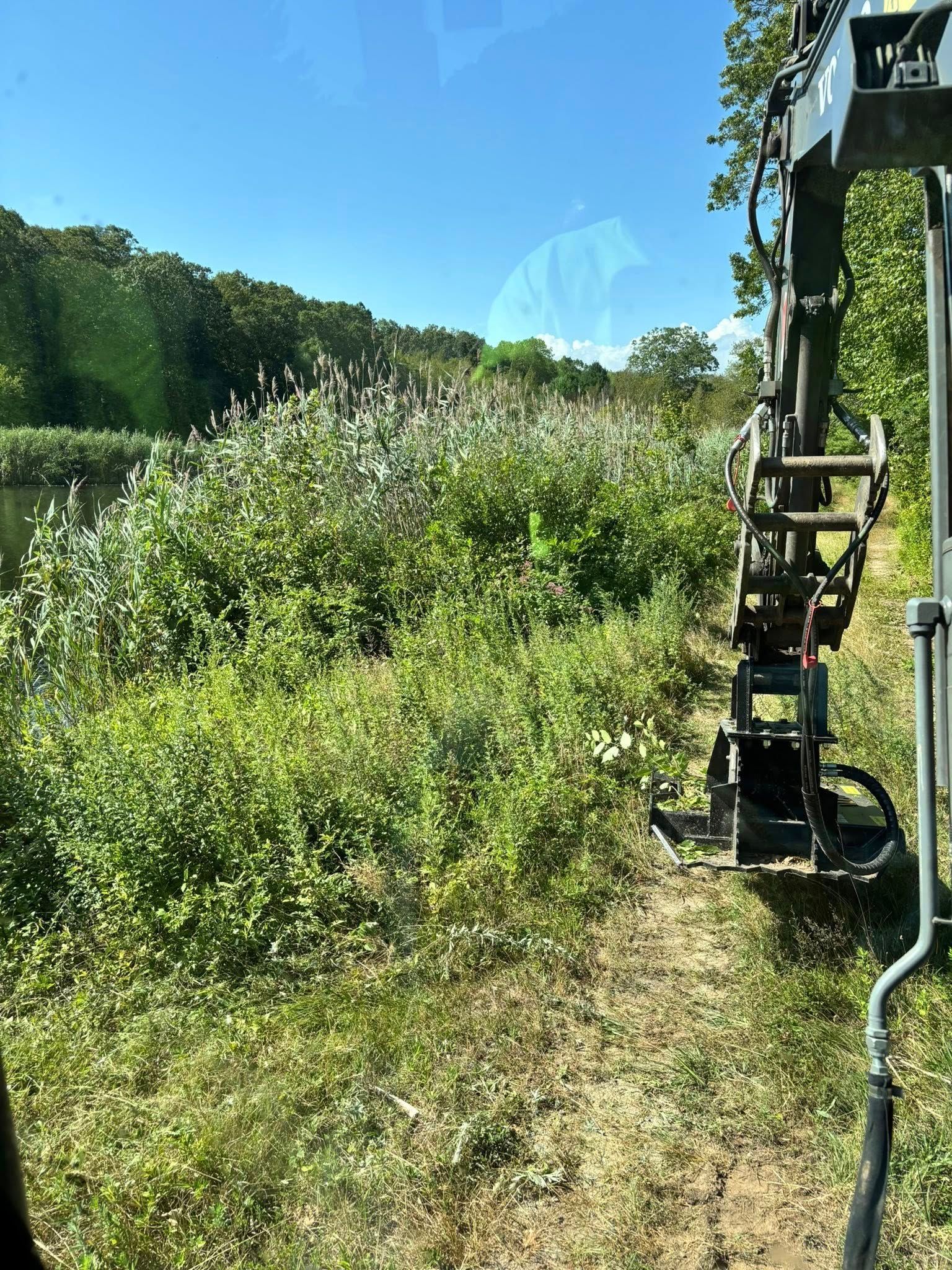 An excavator clearing vegetation along a waterway under a blue sky.