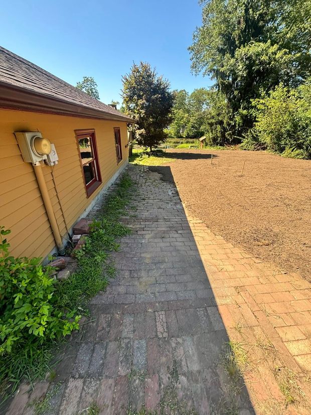 Yellow house exterior with brick path and prepared garden bed in sunlight.
