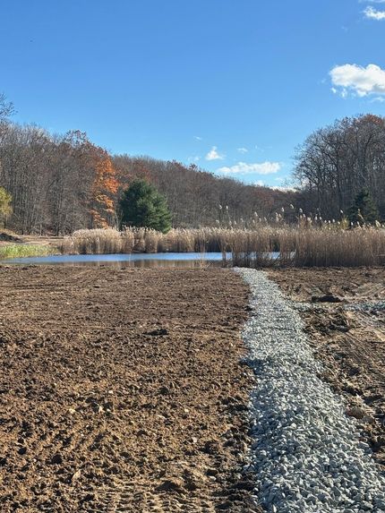 A pond with a gravel path, surrounded by bare earth and trees under a blue sky.