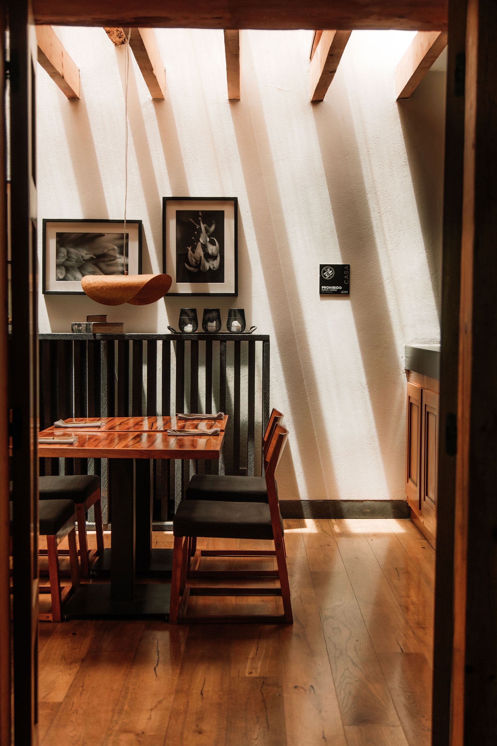 Rincón de comedor soleado con mesa de madera, sillas, cuadros enmarcados y paredes con sombras a rayas.
