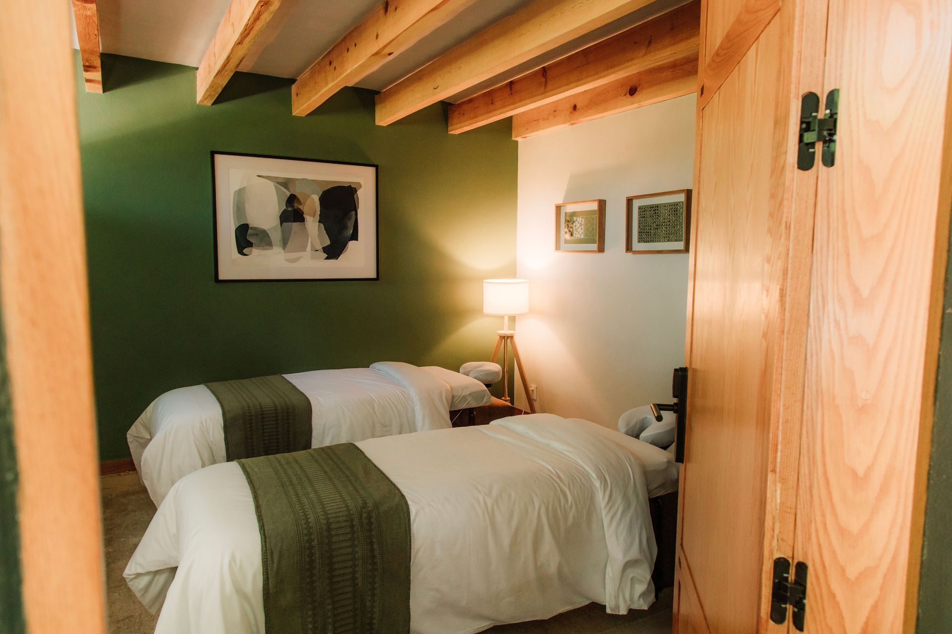 Cabina de masaje acogedora con dos camas individuales, pared decorativa verde, vigas de madera a la vista y lámpara de noche.