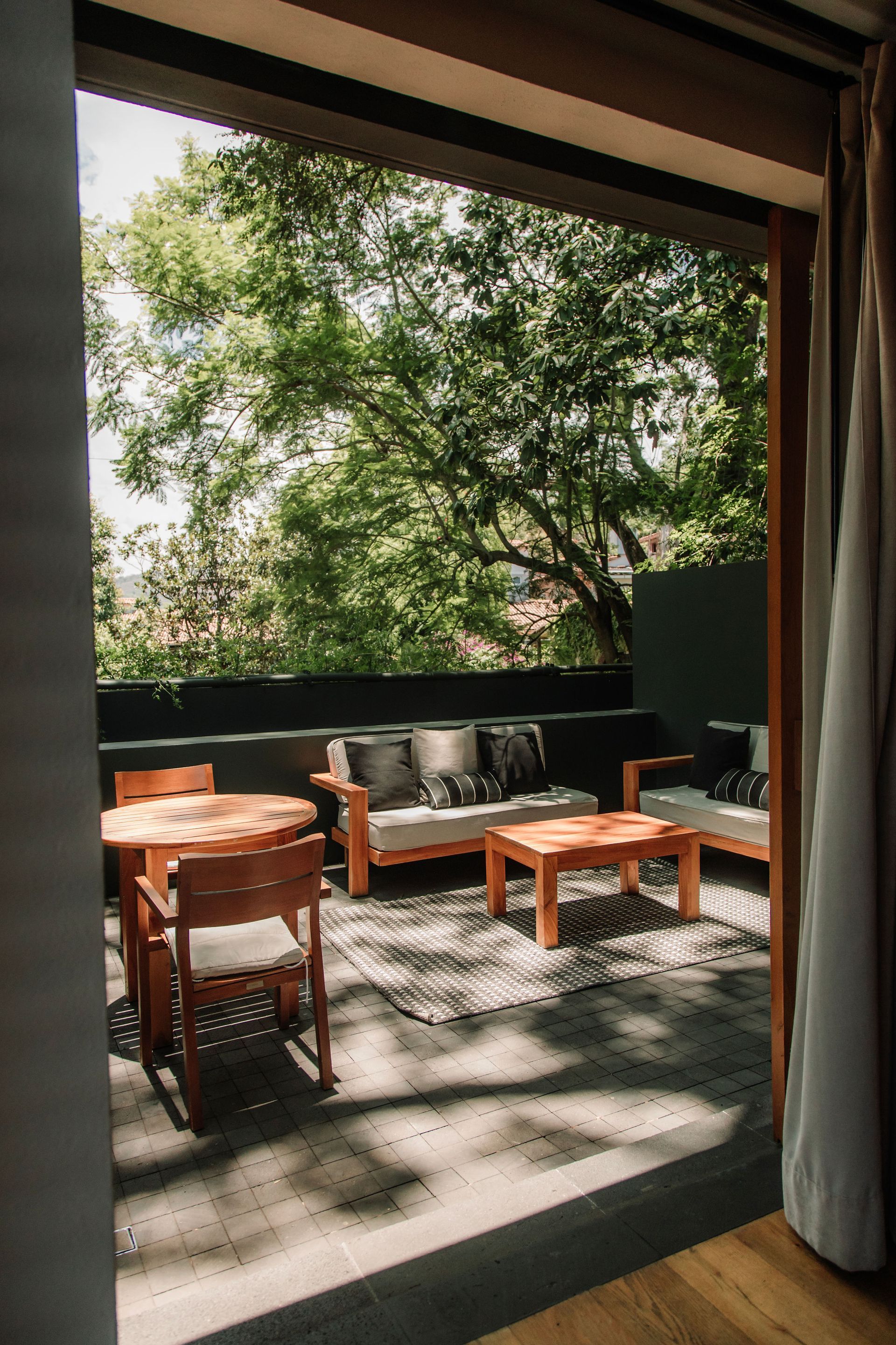Patio cubierto con muebles de madera y plantas en macetas, visible a través de cortinas abiertas, con árboles al fondo.