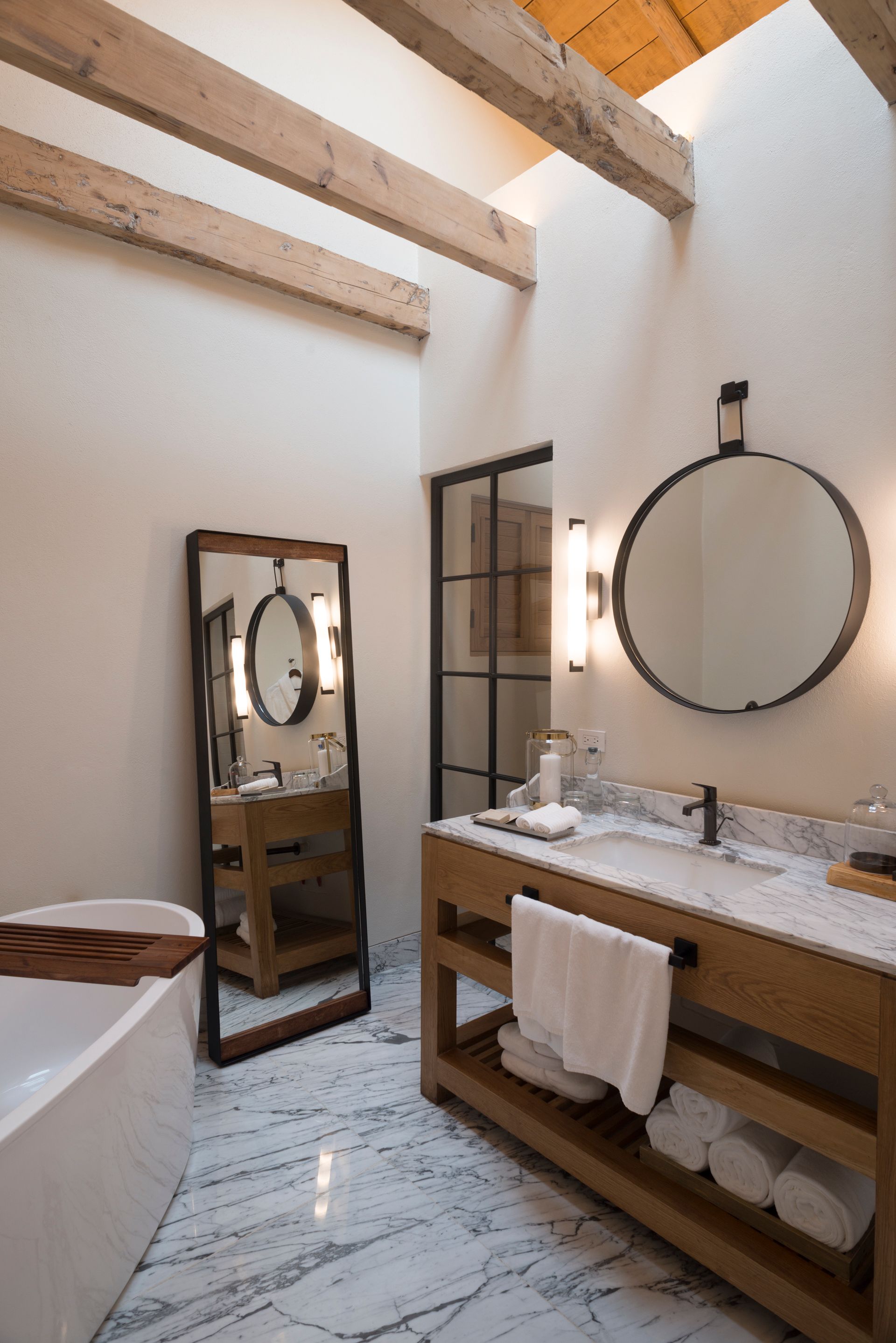 Baño luminoso con techo de vigas de madera, espejo grande, lavabo doble y espejo de pared redondo.