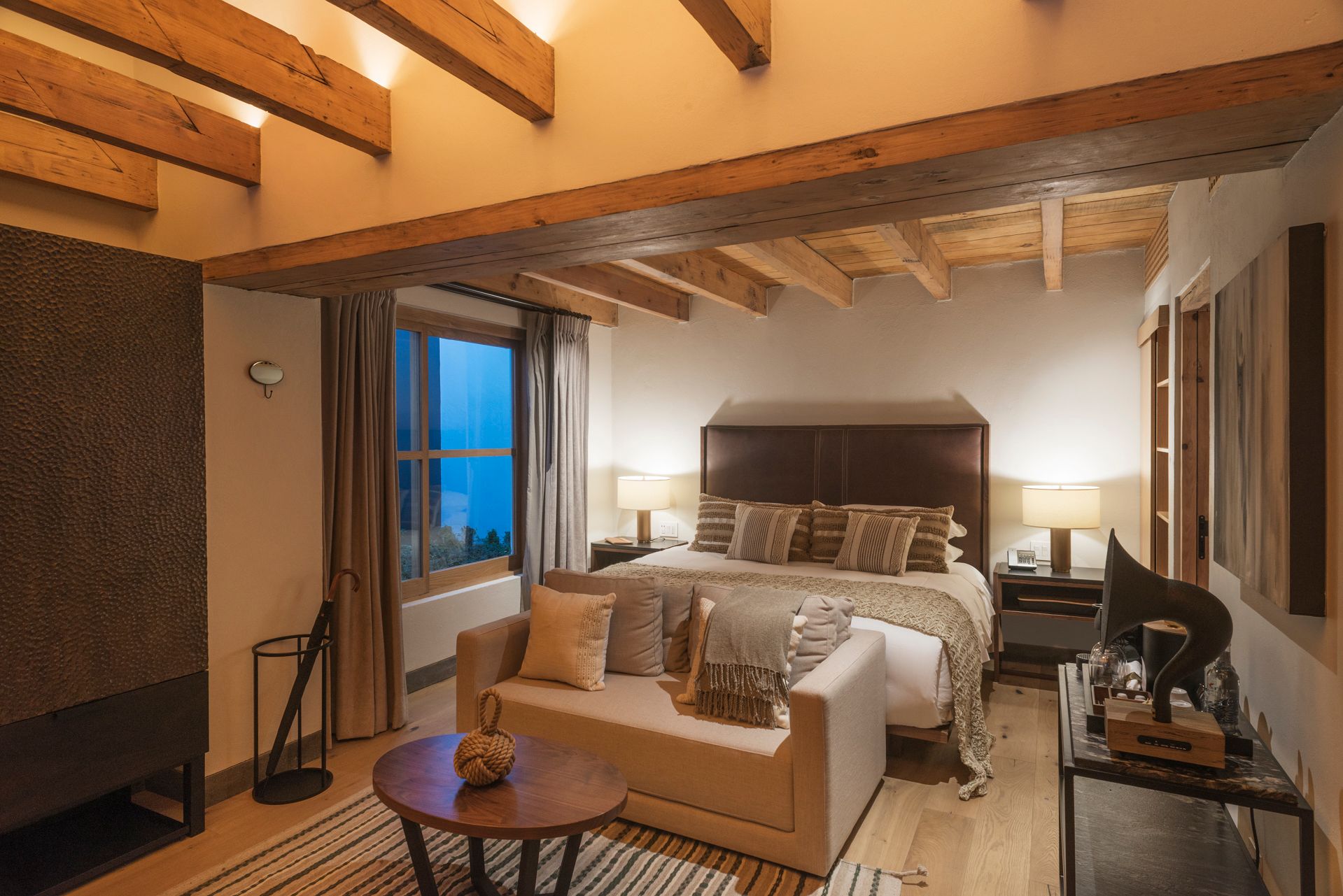 Habitación acogedora con vigas de madera, cama grande, sofá y luz ambiental cálida.