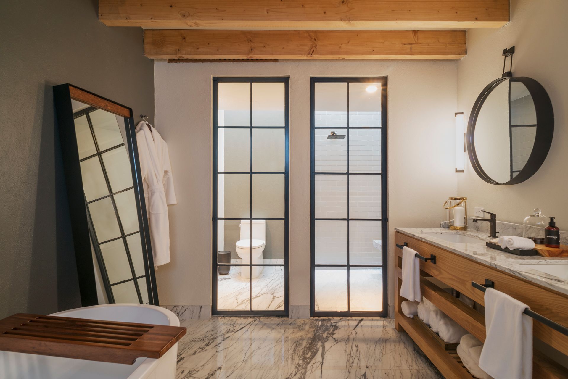 Baño moderno con bañera exenta, tocador de madera, espejo redondo y mampara de ducha de cristal.