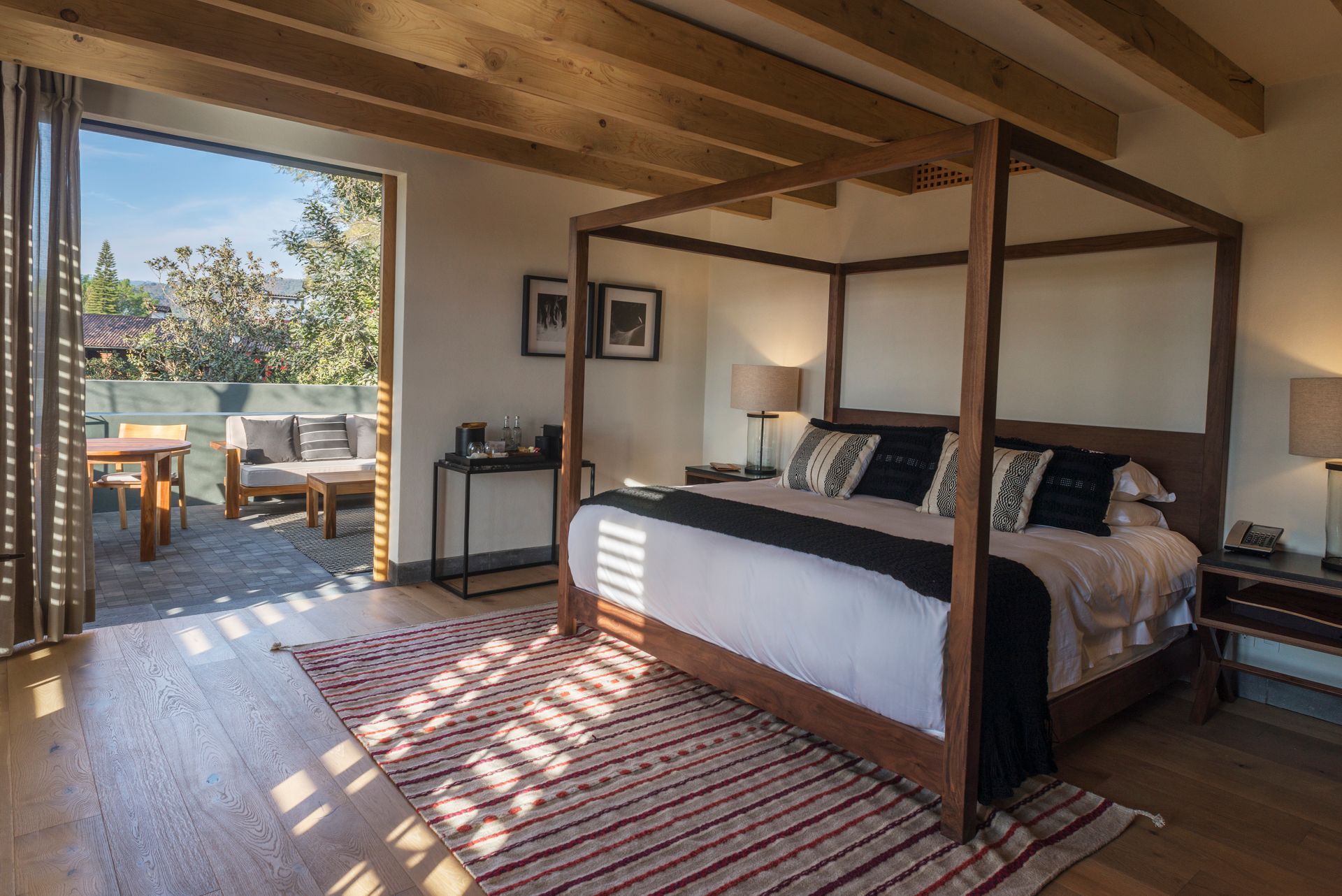 Habitación luminosa con cama con dosel, vigas de madera y puertas que dan a un patio soleado.