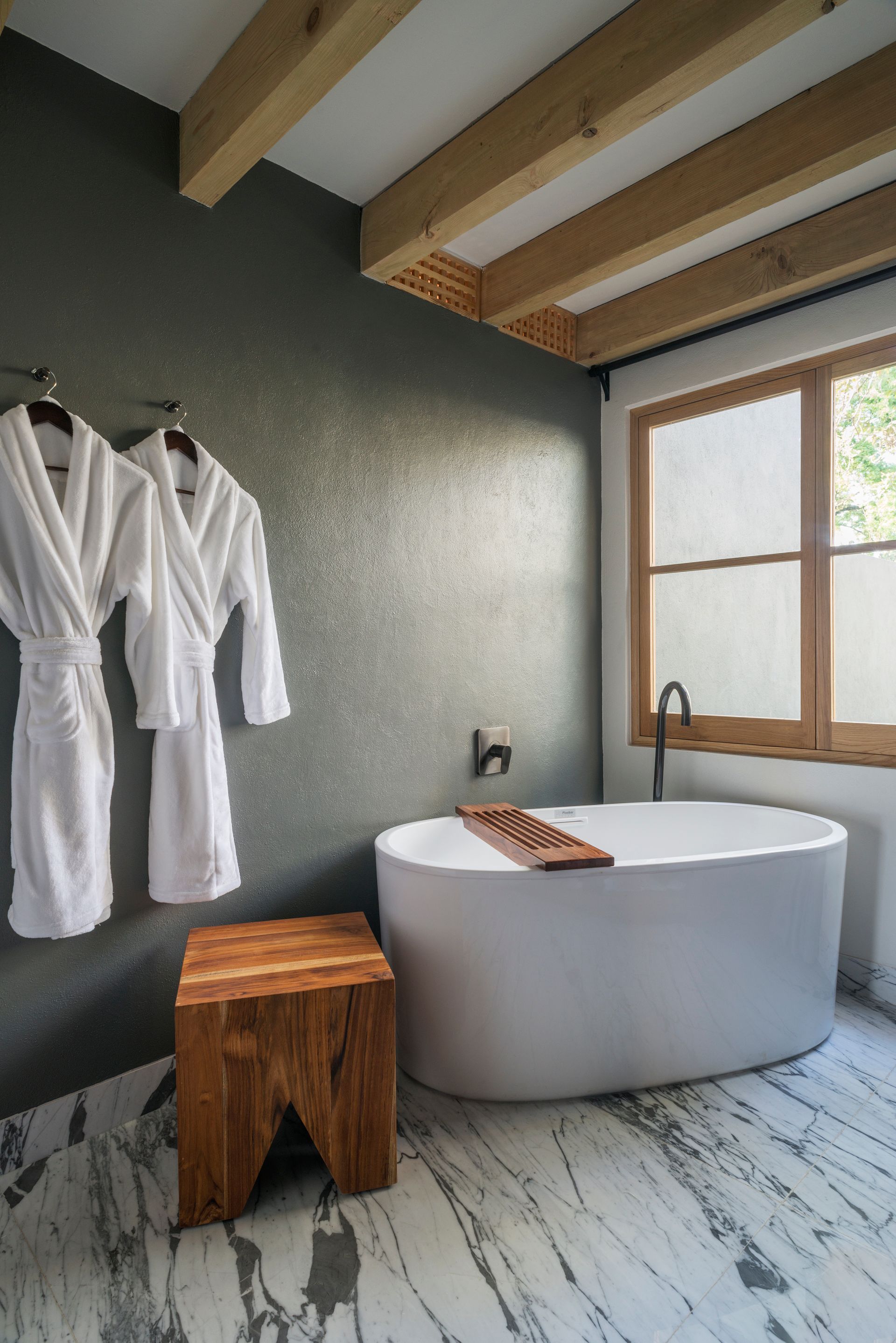 Baño moderno con bañera redonda, taburete de madera, albornoces y ventana en un entorno de spa rústico.