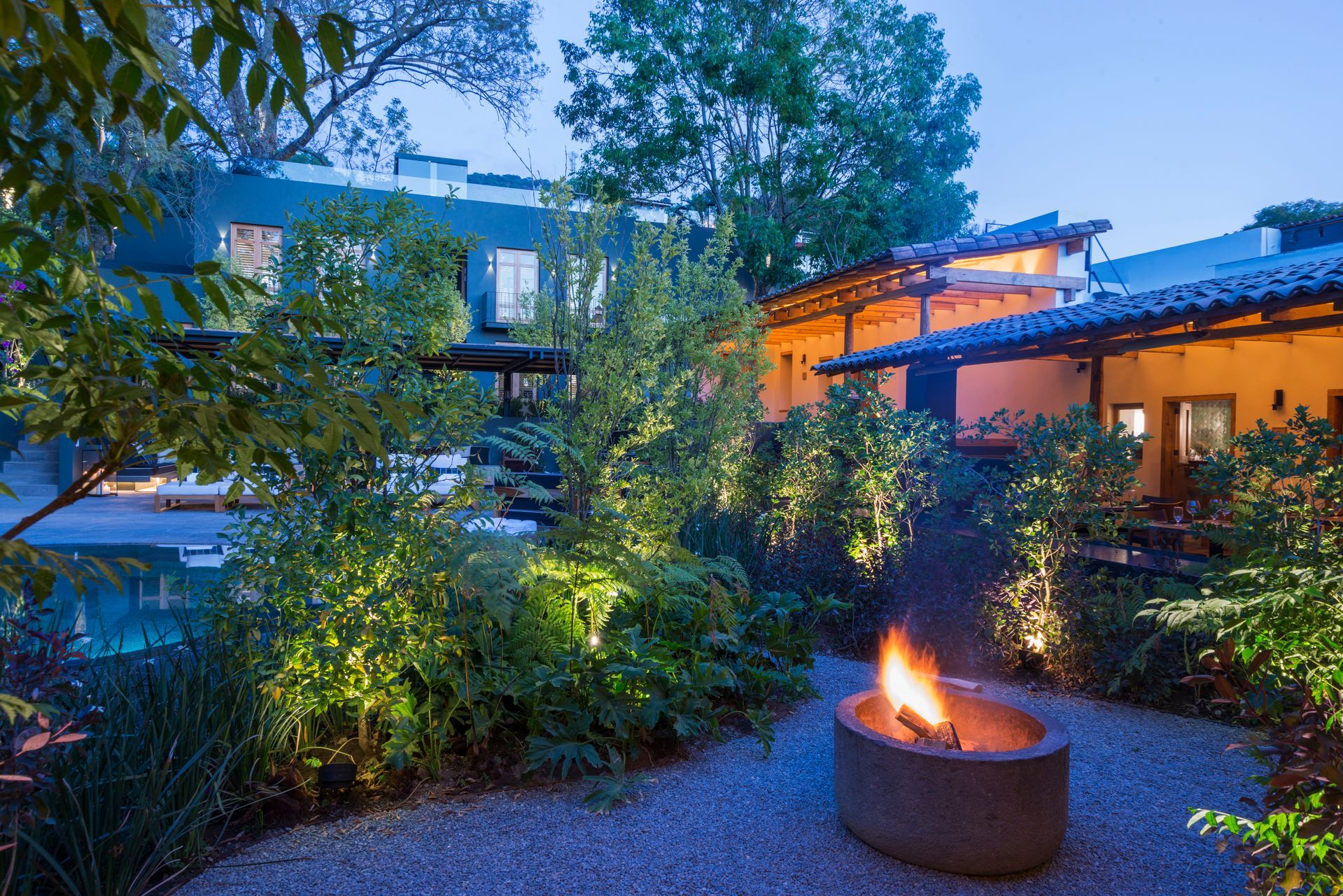 Patio ajardinado al atardecer con una hoguera encendida, exuberante vegetación y casas modernas al fondo.