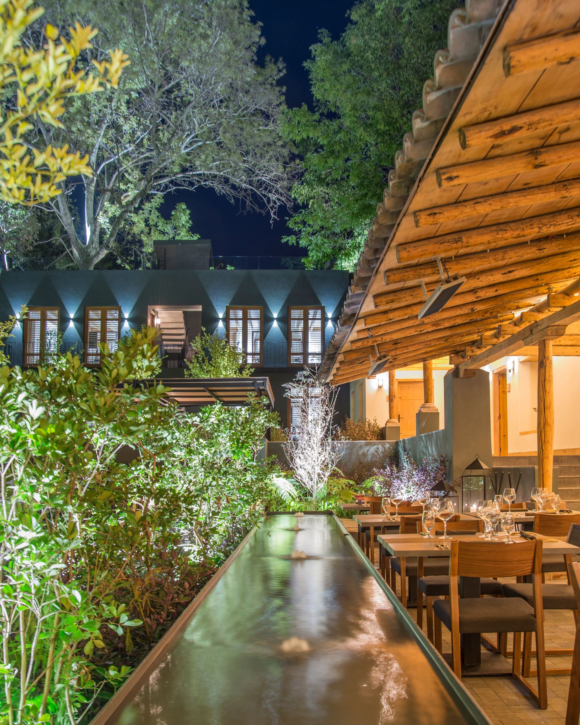 Terraza de restaurante al aire libre por la noche, con iluminación cálida, mesas de madera, vegetación y árboles.