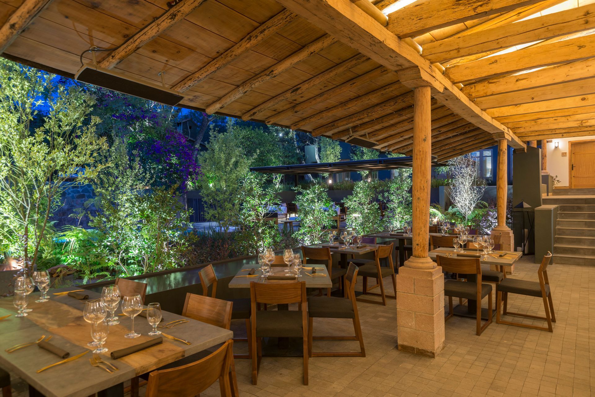 Acogedor patio exterior de restaurante con mesas y sillas de madera, iluminación cálida y exuberante vegetación al atardecer.