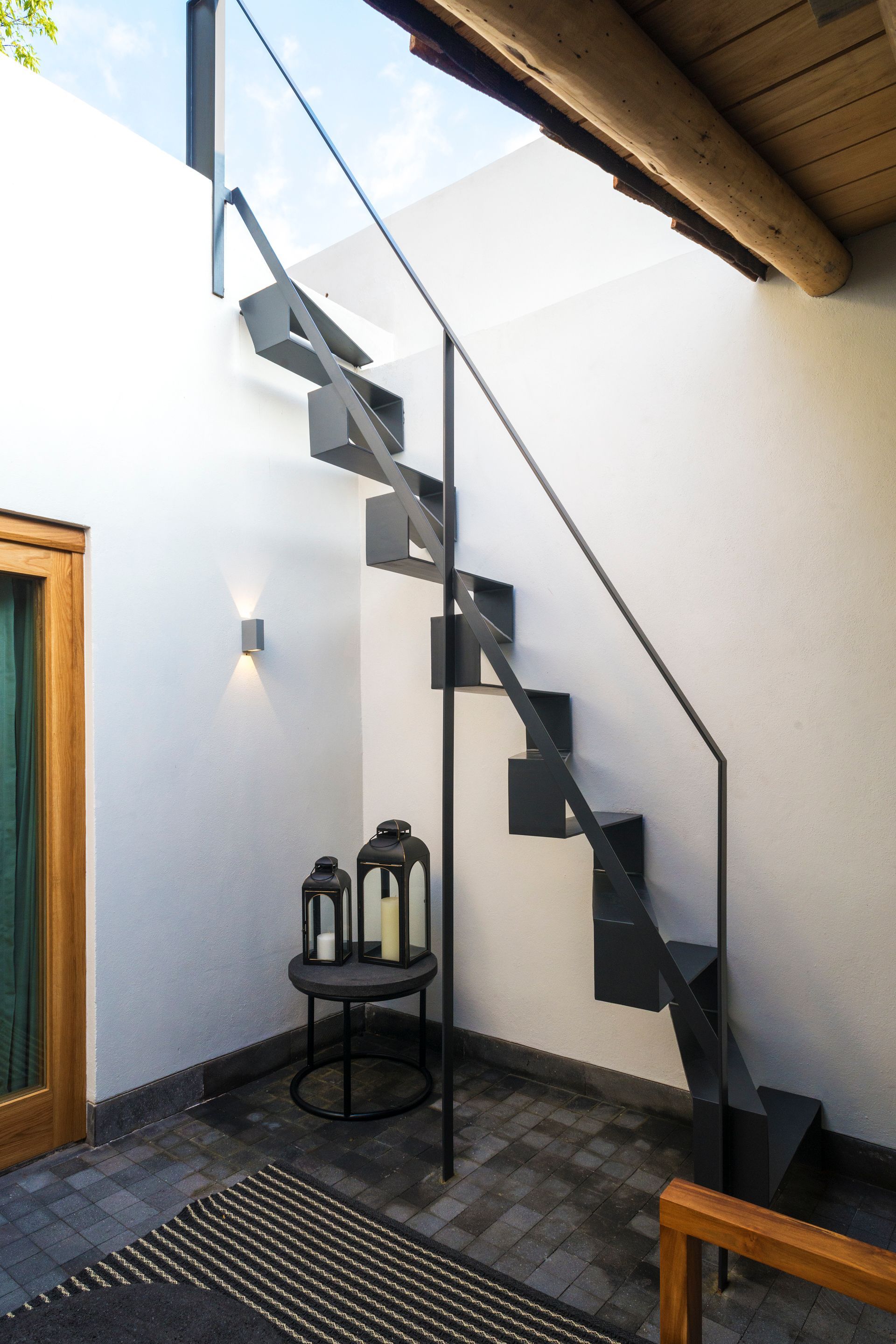 Escalera exterior con peldaños y barandilla de metal oscuro junto a una pared blanca en un patio.