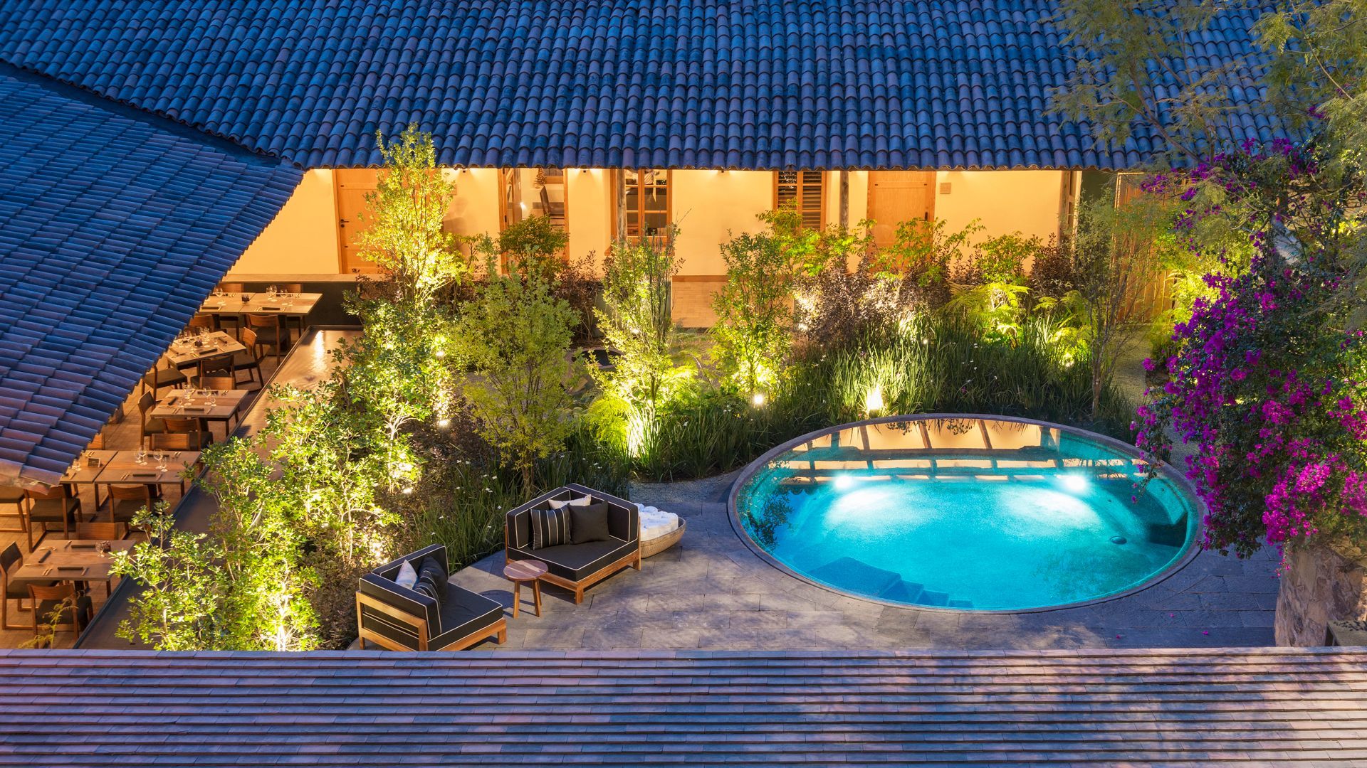 Piscina iluminada en el patio al anochecer, con tumbonas, plantas exuberantes y luces cálidas junto a un edificio.