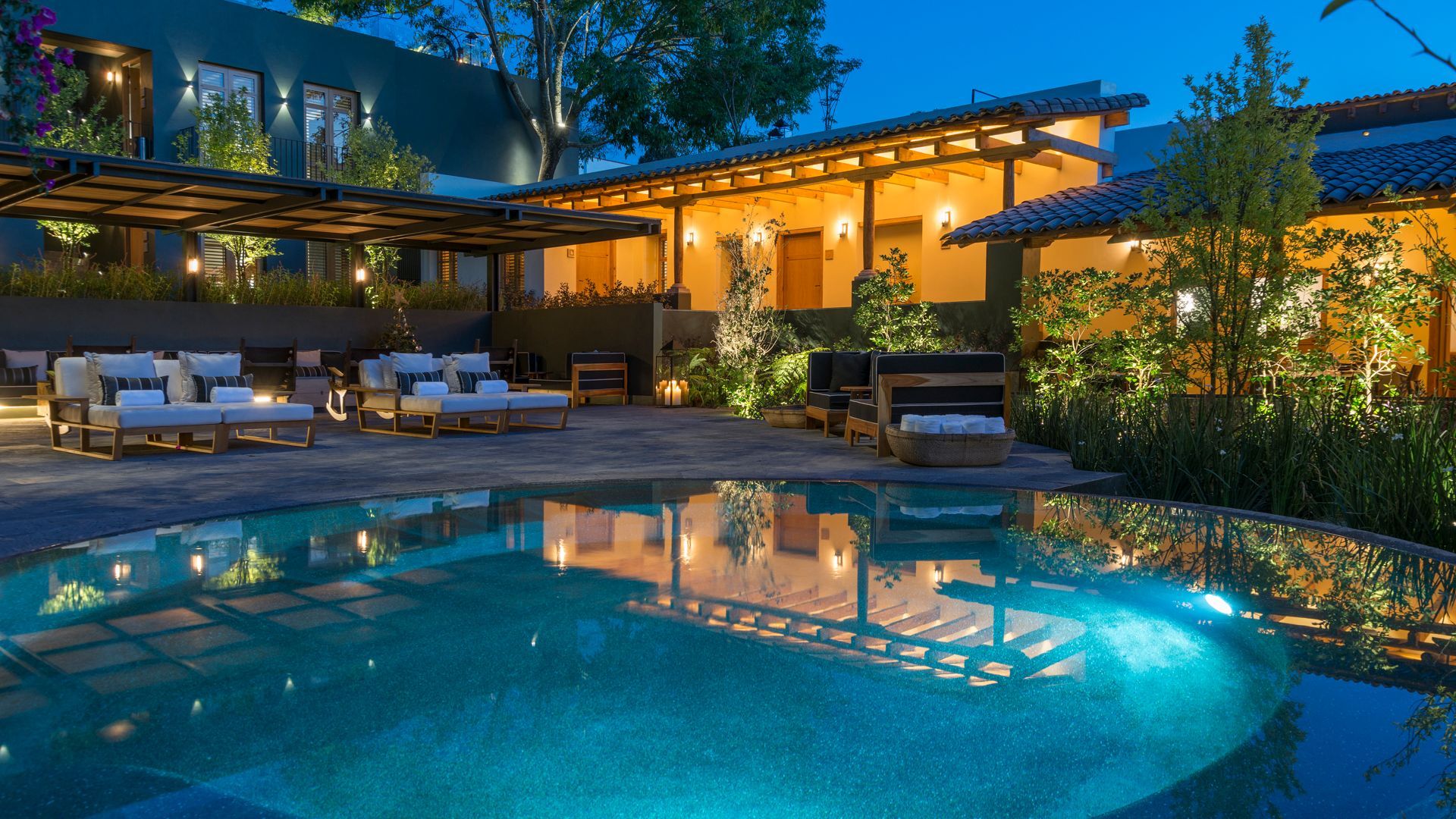 Lujoso patio trasero con piscina iluminada, tumbonas y una cálida casa moderna al atardecer.