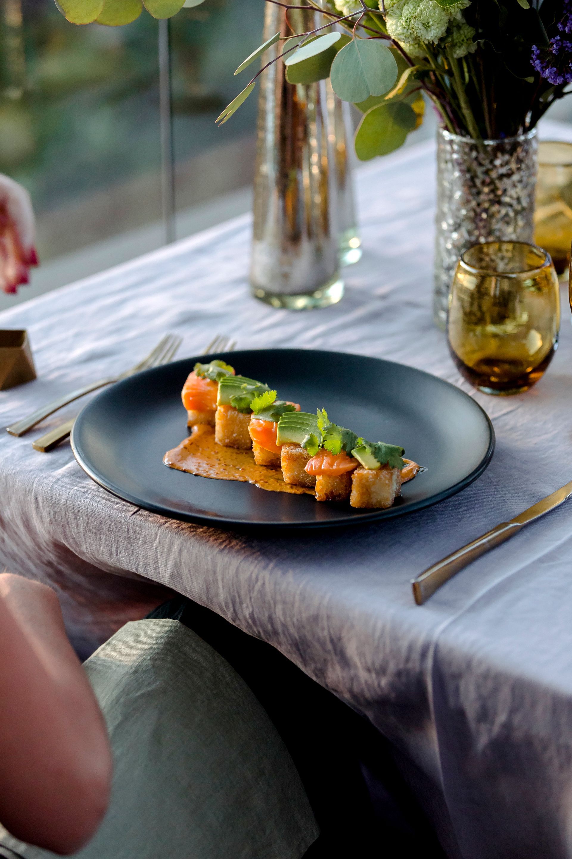 Aperitivo emplatado con salmón y verduras sobre un mantel gris junto a un jarrón y cristalería junto a una ventana.