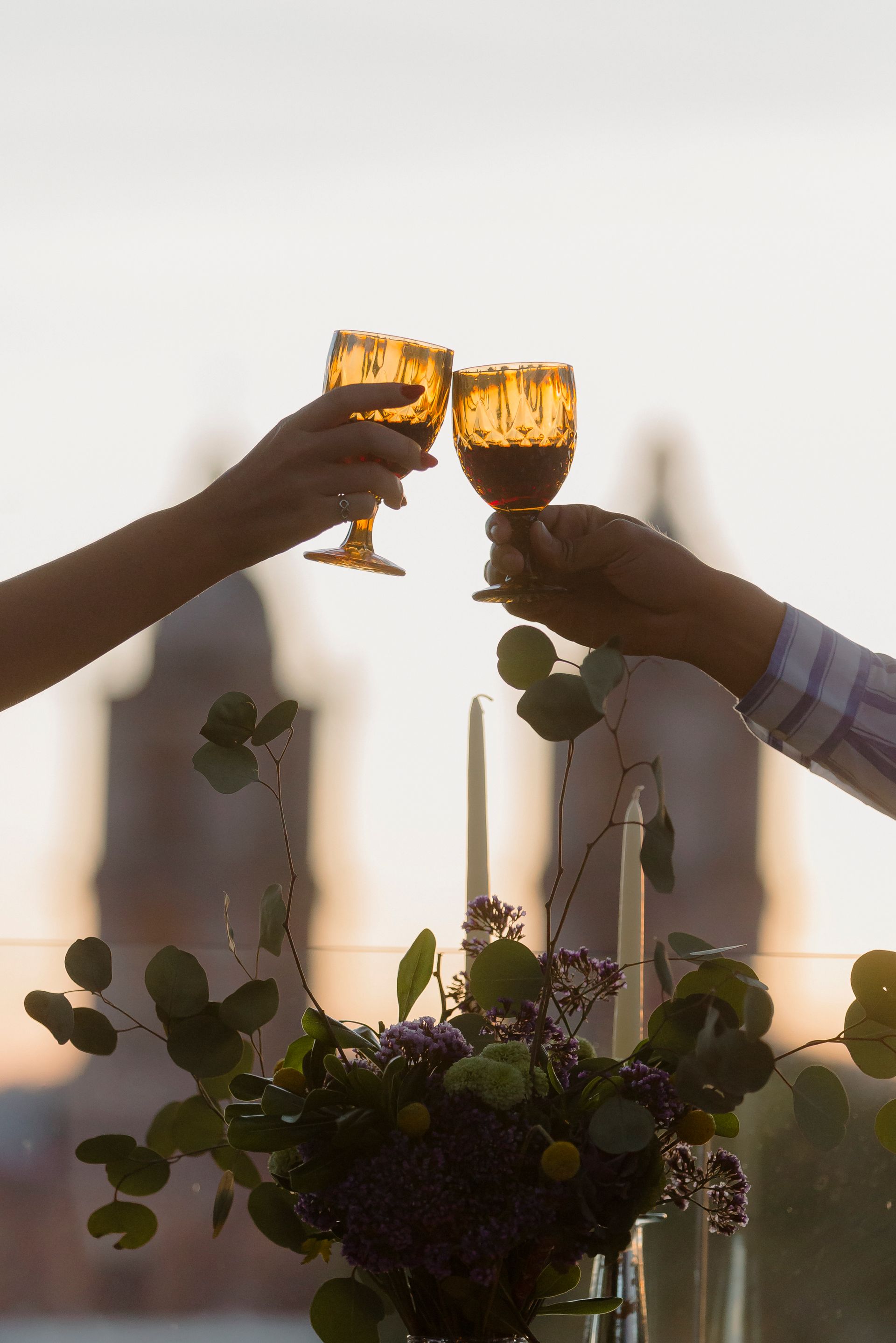 Manos alzando copas de vino ámbar para brindar junto a un centro de mesa floral iluminado al atardecer.