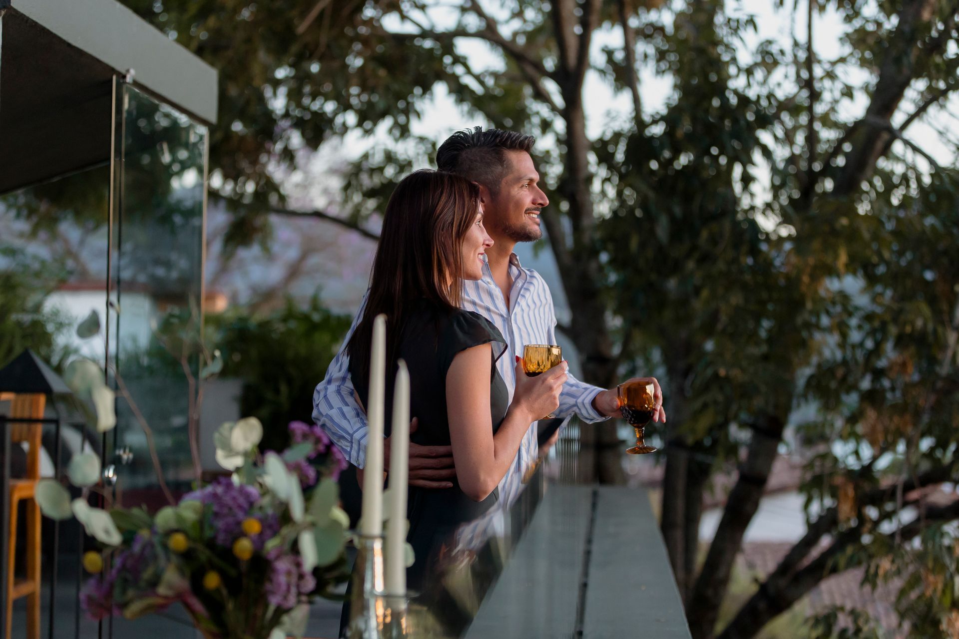 Dos personas con bebidas en la mano en un balcón, contemplando los árboles al atardecer.