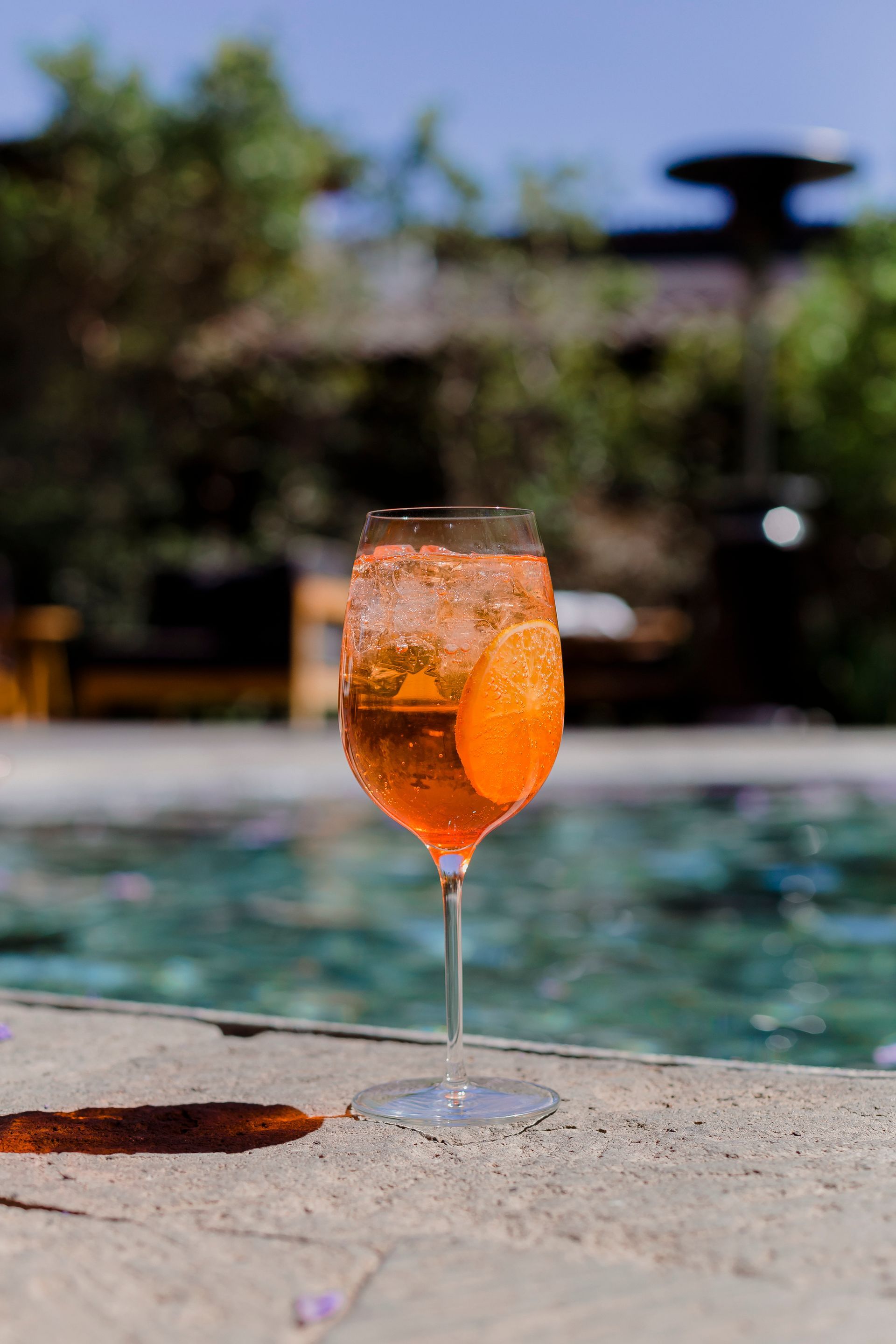 Cóctel de naranja en una copa de tallo junto a una piscina en un patio soleado.
