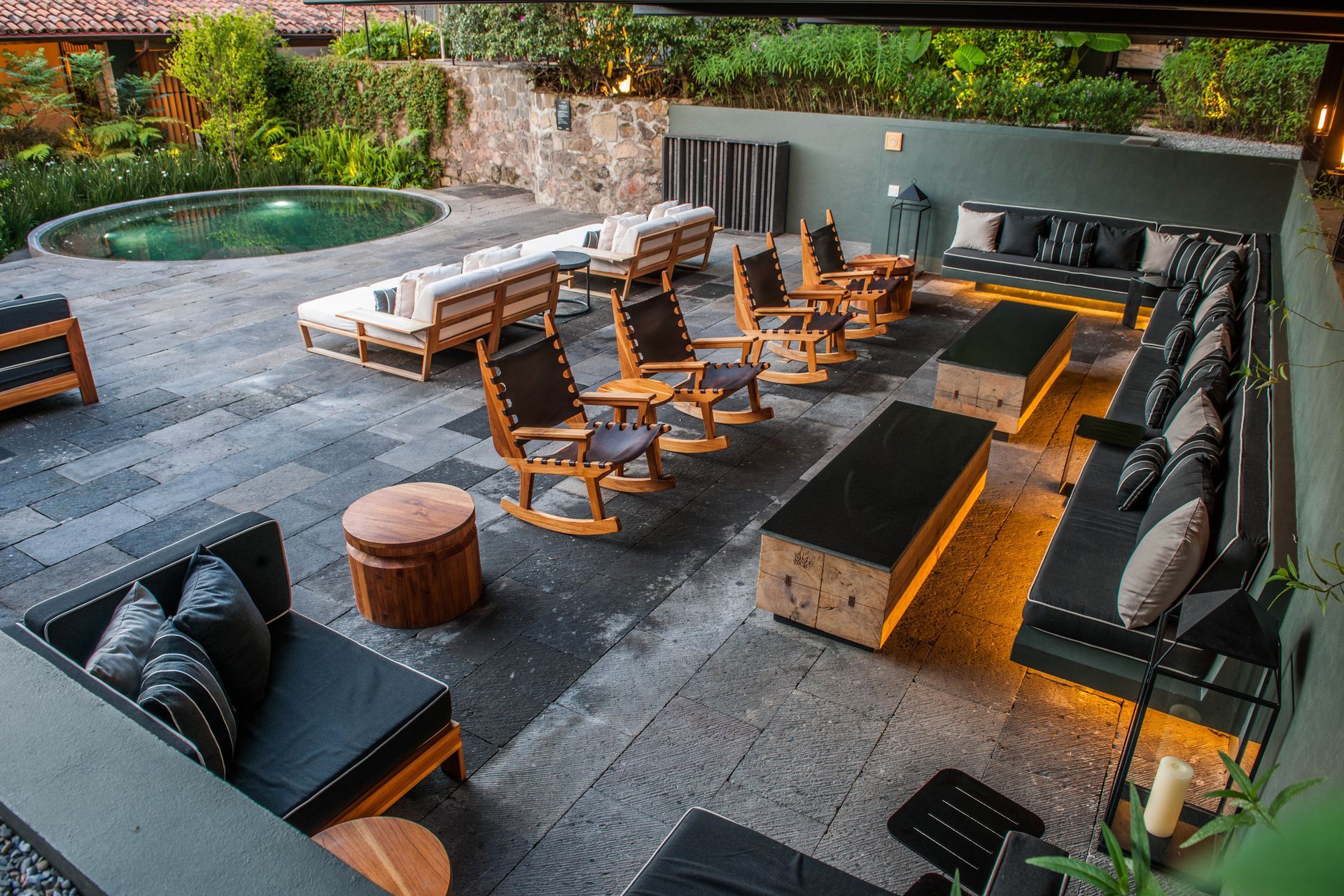 Salón de patio al aire libre con fogata, asientos acolchados, sillas de madera y una pequeña piscina al fondo.