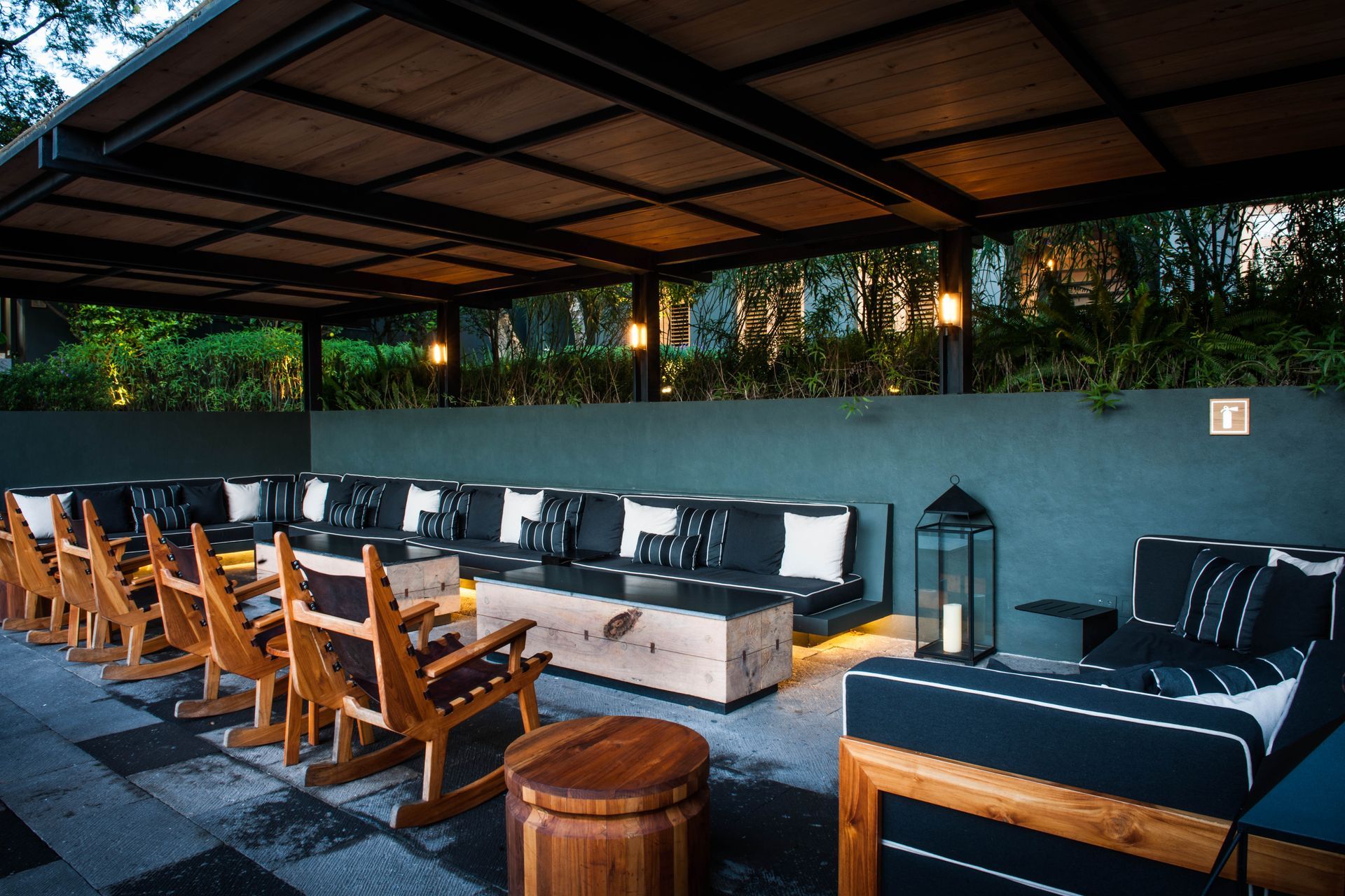 Salón al aire libre con sillas de madera, sofás y faroles bajo un techo cubierto al atardecer.