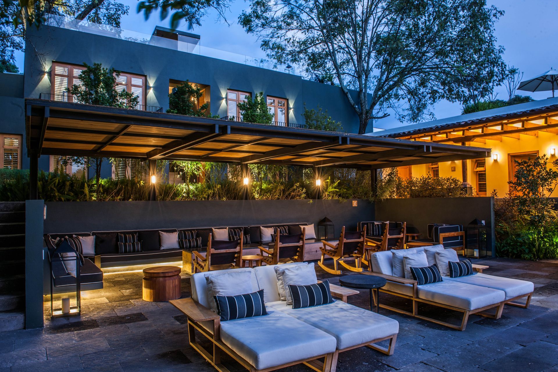 Terraza exterior con tumbonas acolchadas y luces de guirnalda cálidas bajo una pérgola moderna al atardecer.