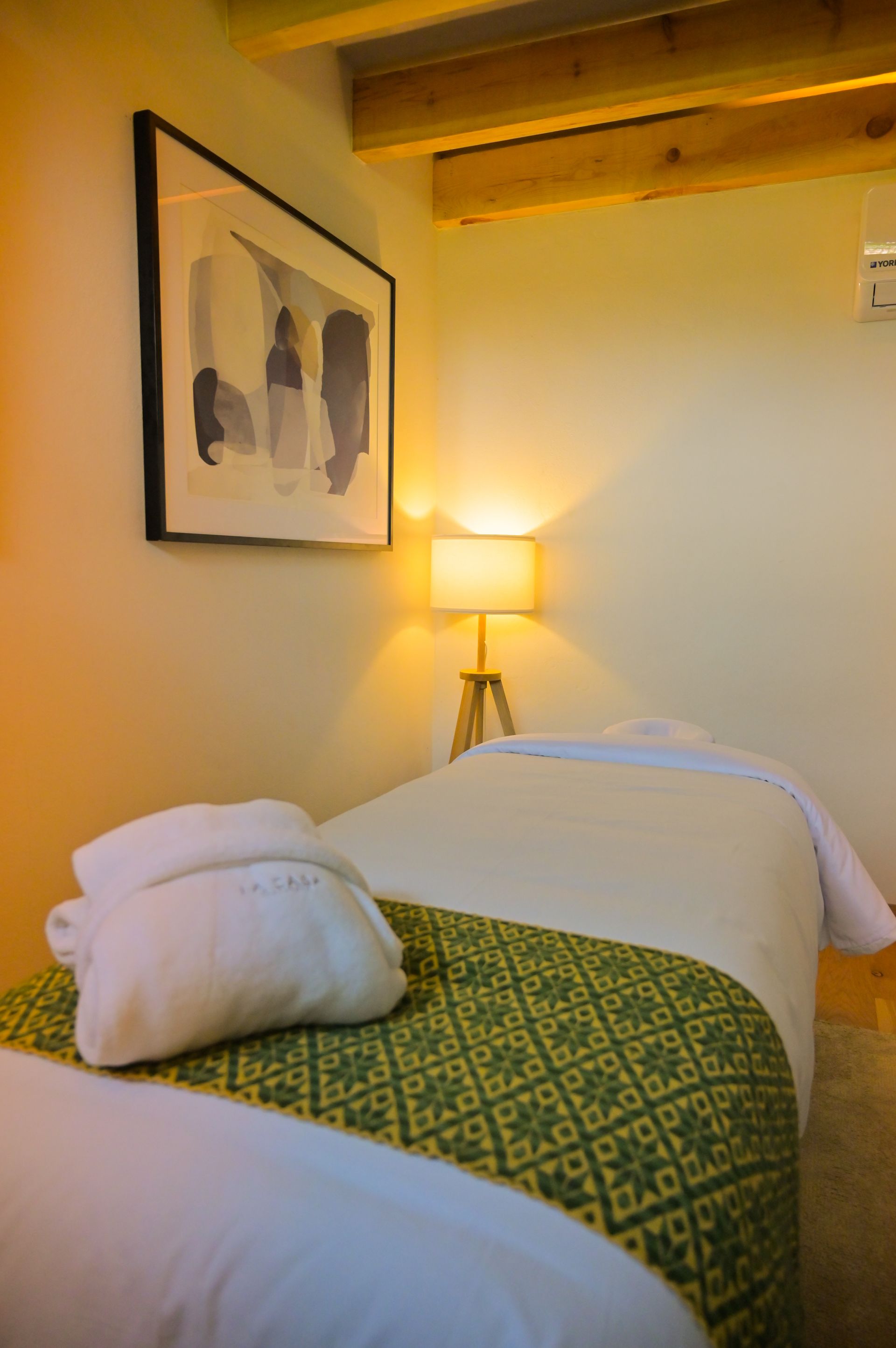 Cabina de masaje  acogedora con cama individual, manta estampada verde, lámpara de noche y cuadro enmarcado.